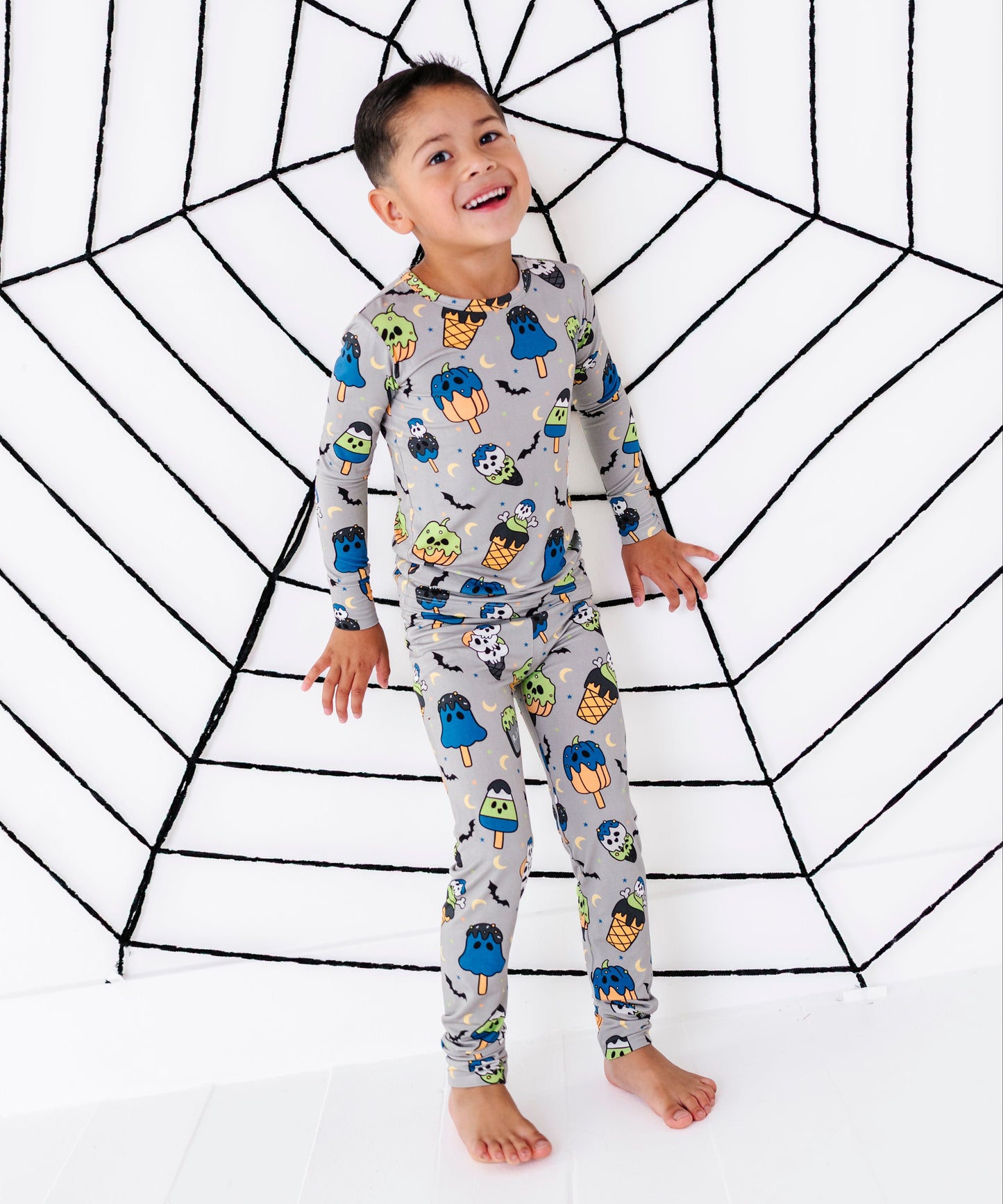 A young boy stands barefoot in front of a white studio backdrop with a large black spiderweb graphic, wearing a light gray long-sleeve pajama set covered in colorful cartoon spider and monster prints.
