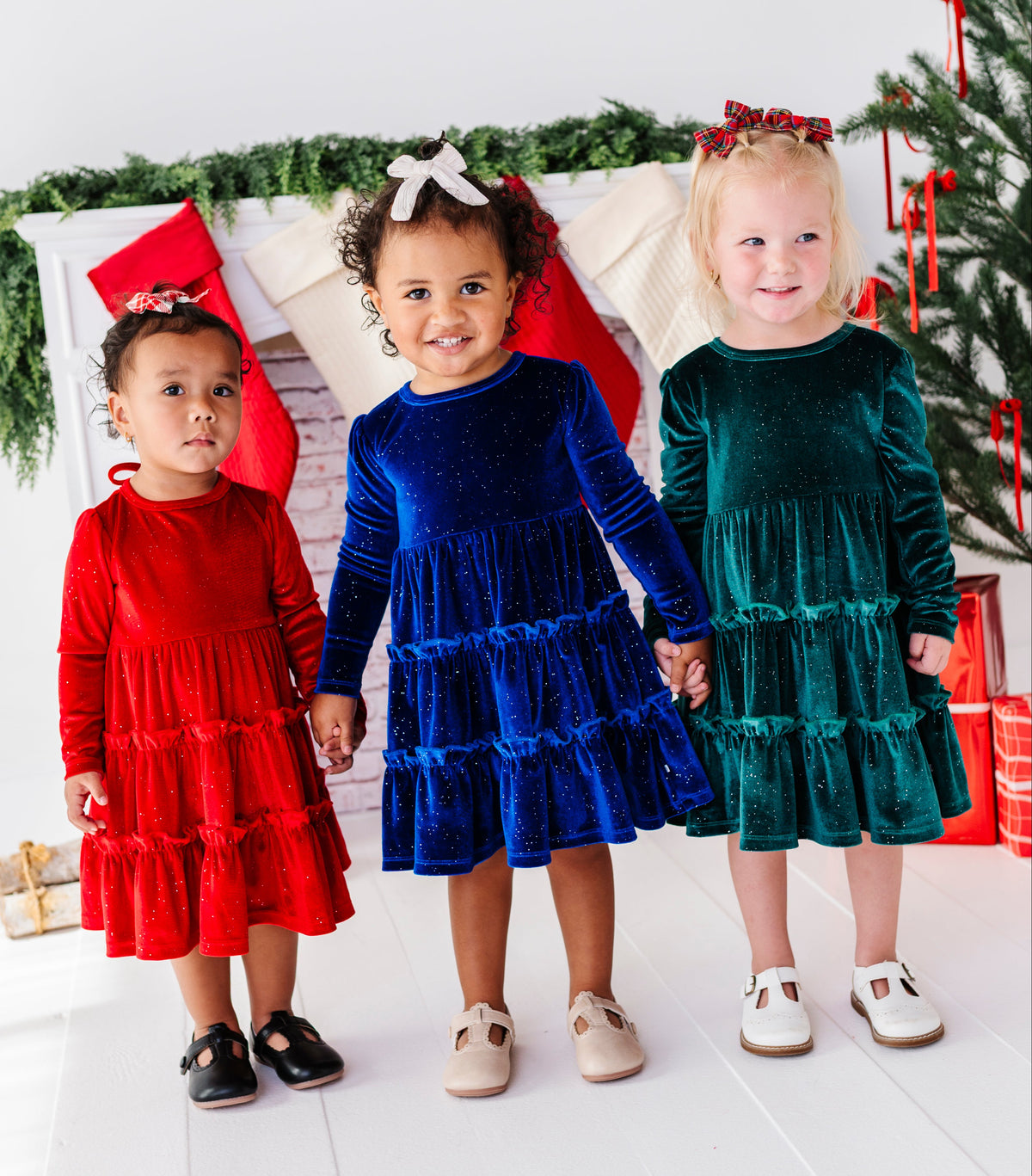 Three young children standing together in festive holiday attire, wearing tiered velvet dresses in red, royal blue, and emerald green, posed in front of a decorated Christmas tree and stockings.
