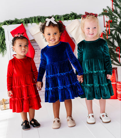 Three young children standing together in festive holiday attire, wearing tiered velvet dresses in red, royal blue, and emerald green, posed in front of a decorated Christmas tree and stockings.