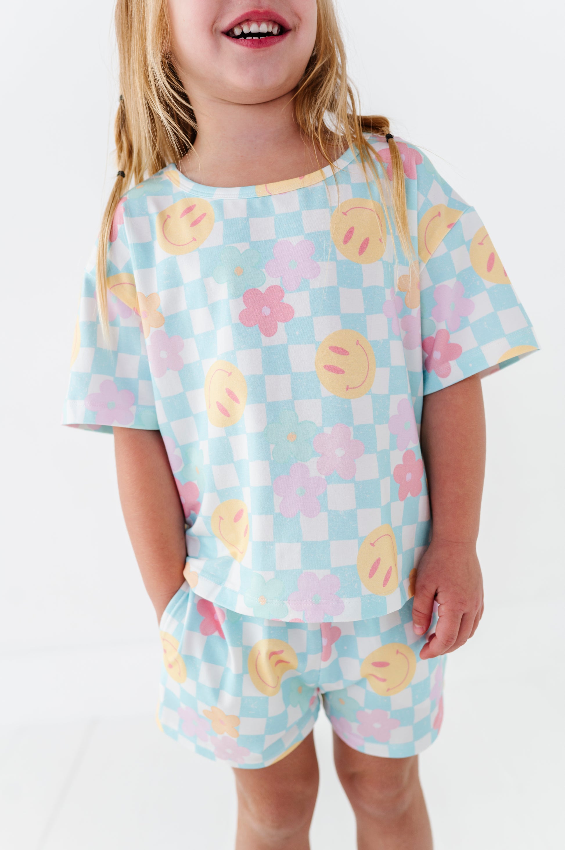 A young girl wearing a matching two-piece pajama set with a pastel checkered pattern and smiley face and flower motifs, photographed against a clean white background.