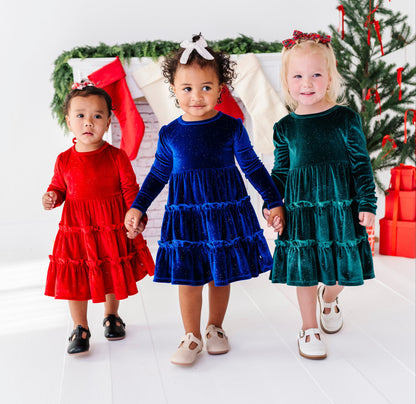 Three young children wearing festive Christmas dresses in red, royal blue, and emerald green, standing together in front of a decorated Christmas tree and stockings.