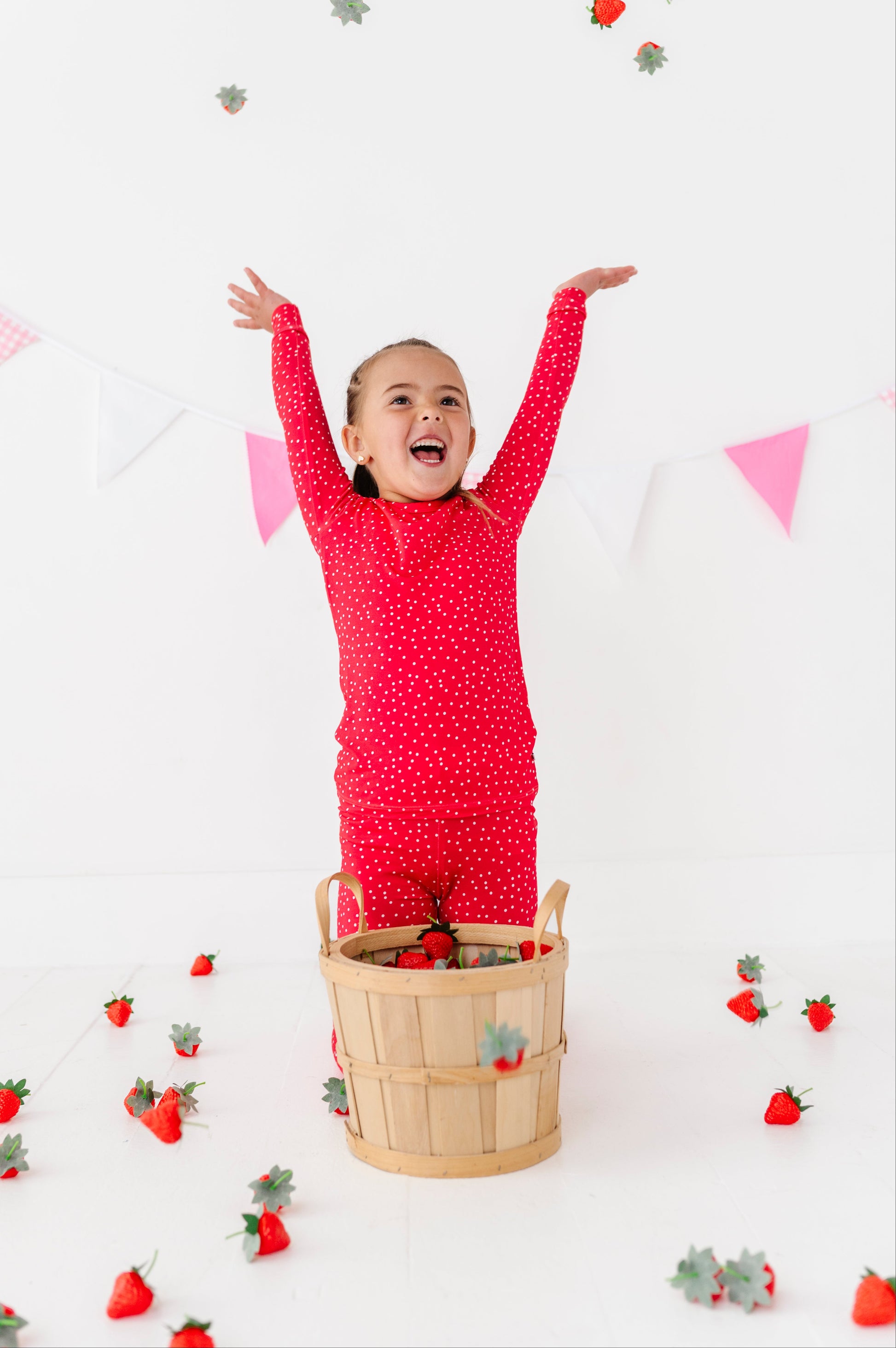 Child in red pajamas with arms raised, standing in a basket surrounded by decorative strawberries on a white background.