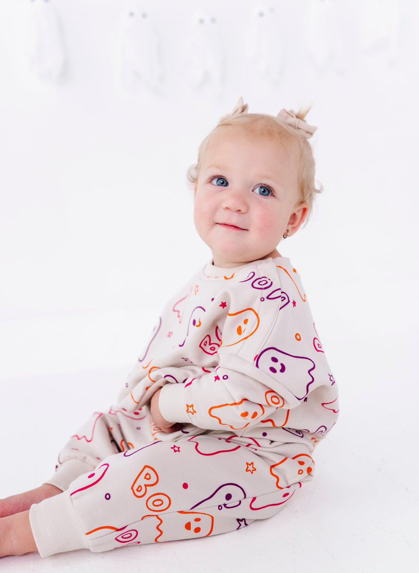 A baby girl sitting on a white floor wearing a cream-colored jumpsuit with a colorful cartoon ghost pattern, looking at the camera.