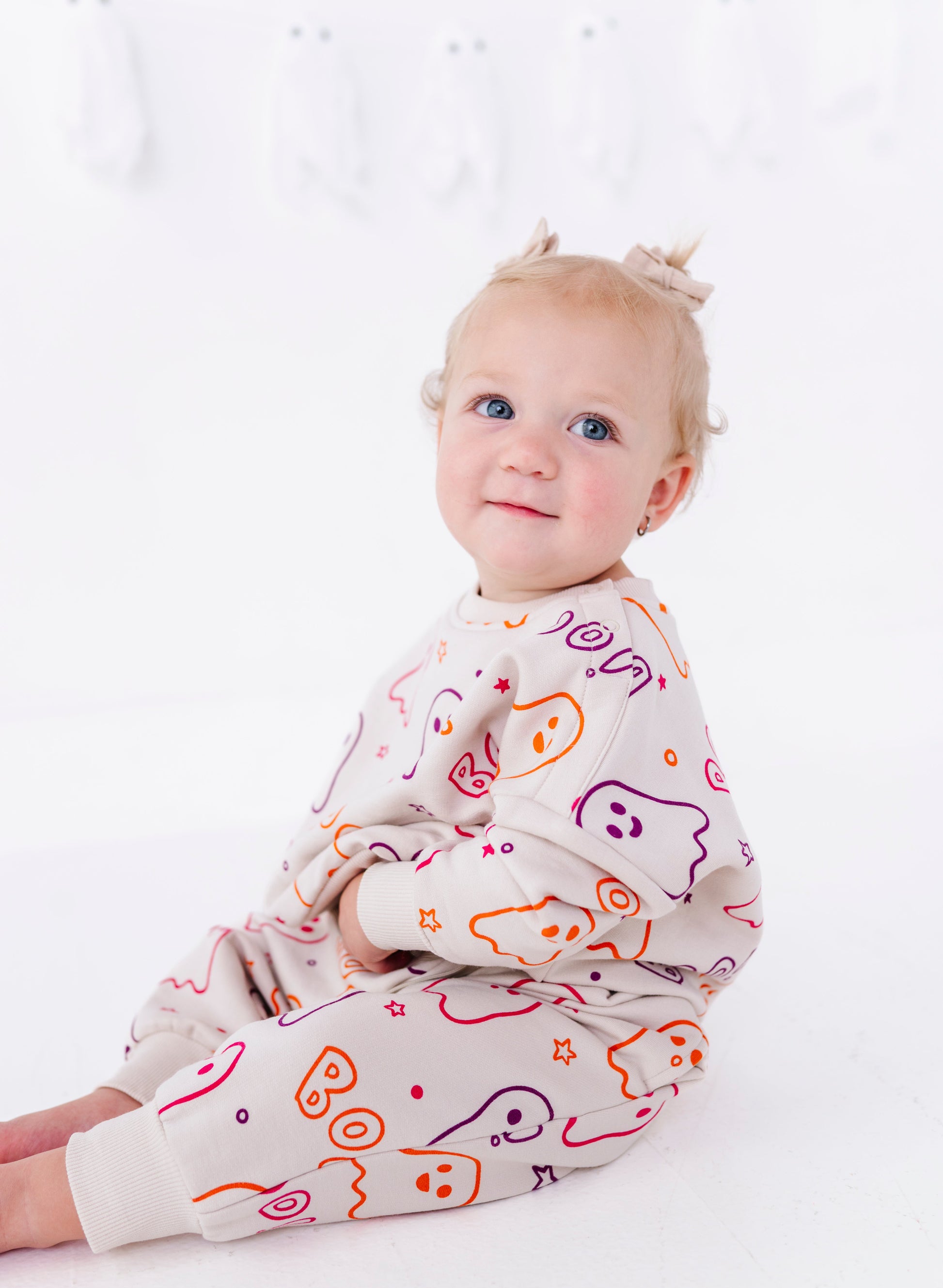 A baby girl sitting on a white floor wearing a cream-colored jumpsuit with a colorful cartoon ghost pattern, looking at the camera.