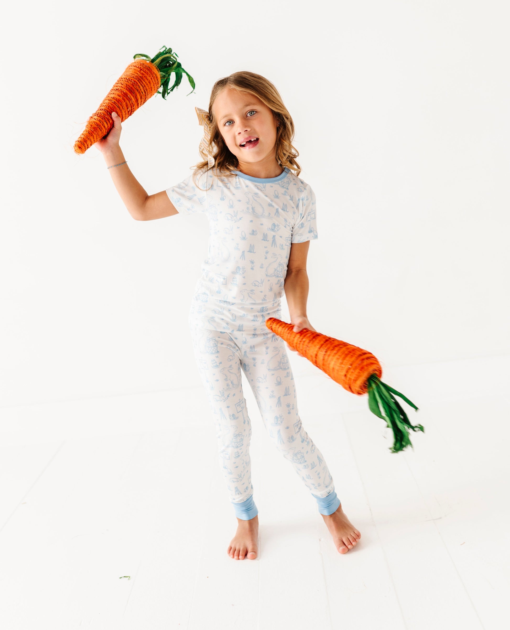 Child wearing a light blue and white Easter pajama set holding two toy carrots on a white background
