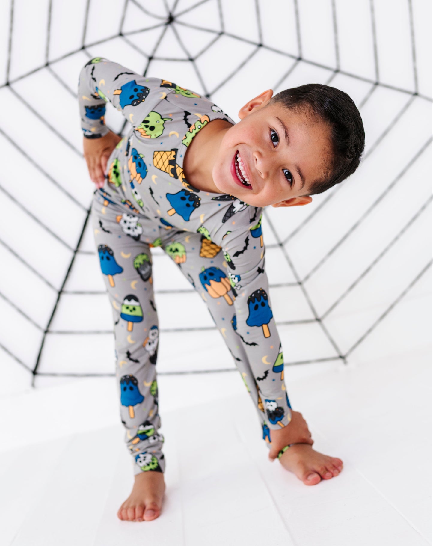 A smiling young boy wearing a long-sleeve gray pajama set with a colorful monster and popsicle print, posing in front of a black spiderweb backdrop on a white studio floor.