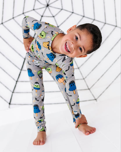 A smiling young boy wearing a long-sleeve gray pajama set with a colorful monster and popsicle print, posing in front of a black spiderweb backdrop on a white studio floor.