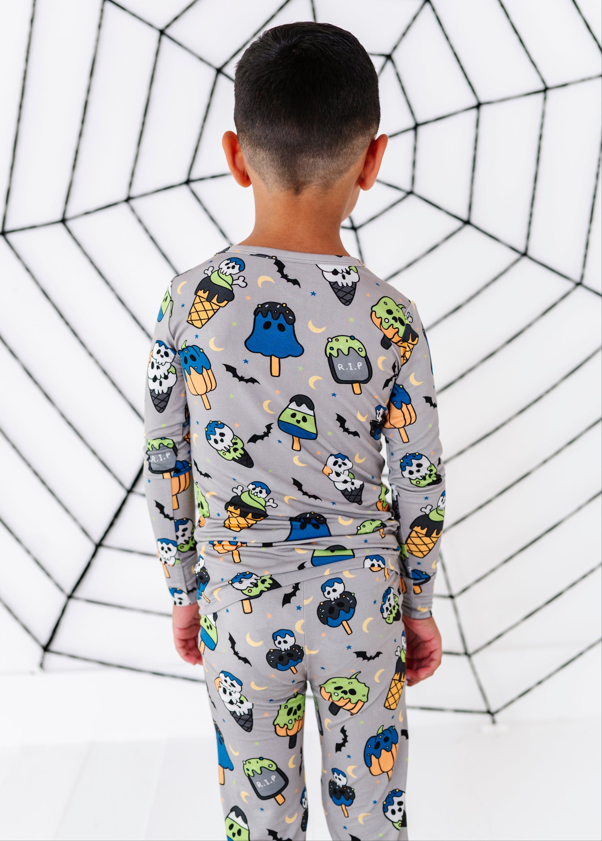 A child viewed from behind wearing a light gray long-sleeve pajama set with a colorful Halloween-themed print of ghosts, bats, pumpkins, and popsicles, standing against a white background with a black spiderweb pattern.