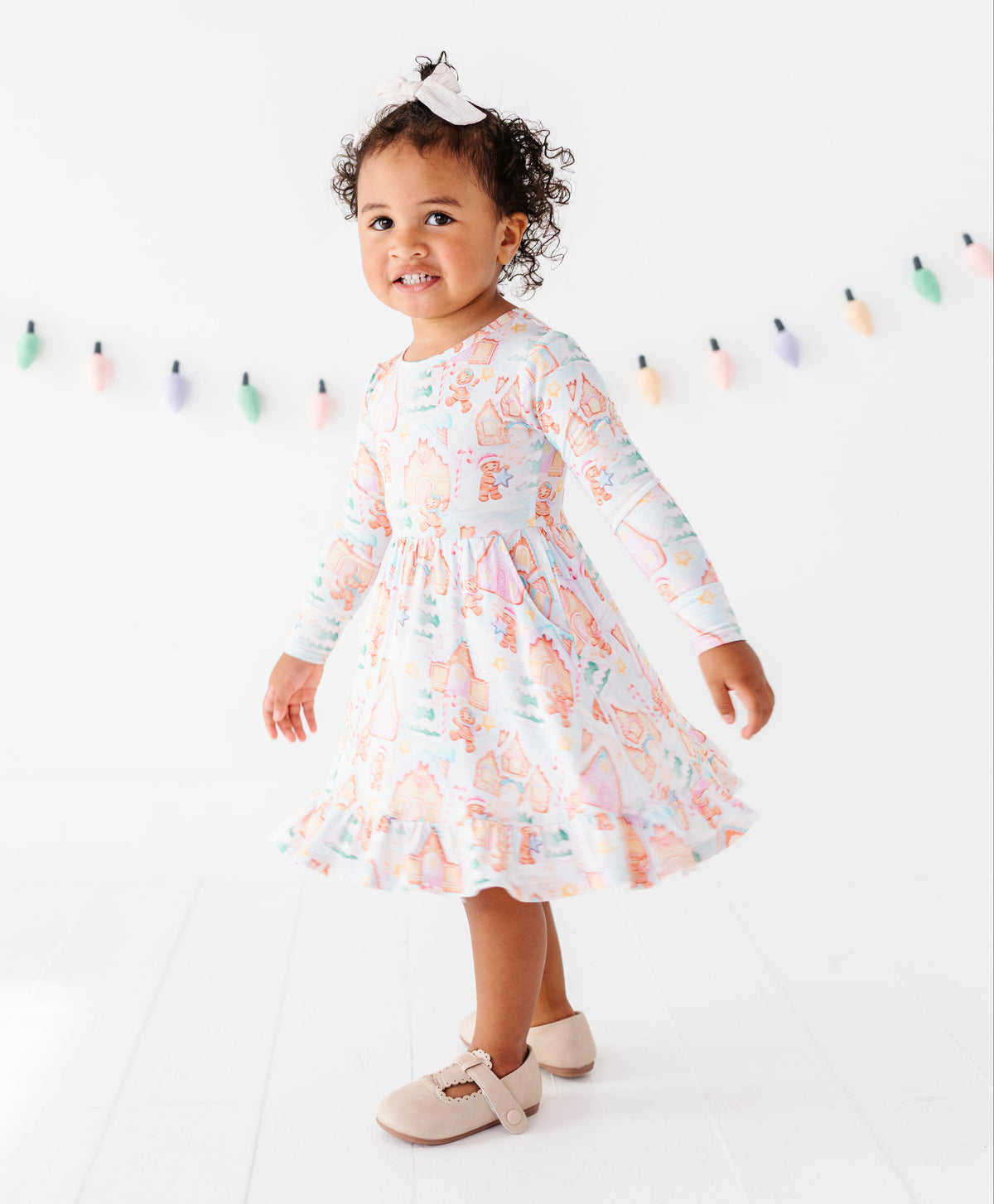 A toddler girl wearing a long-sleeve ruffled dress with a pastel multicolor cartoon print, standing on a white studio floor with a string of colorful hanging lights in the background.