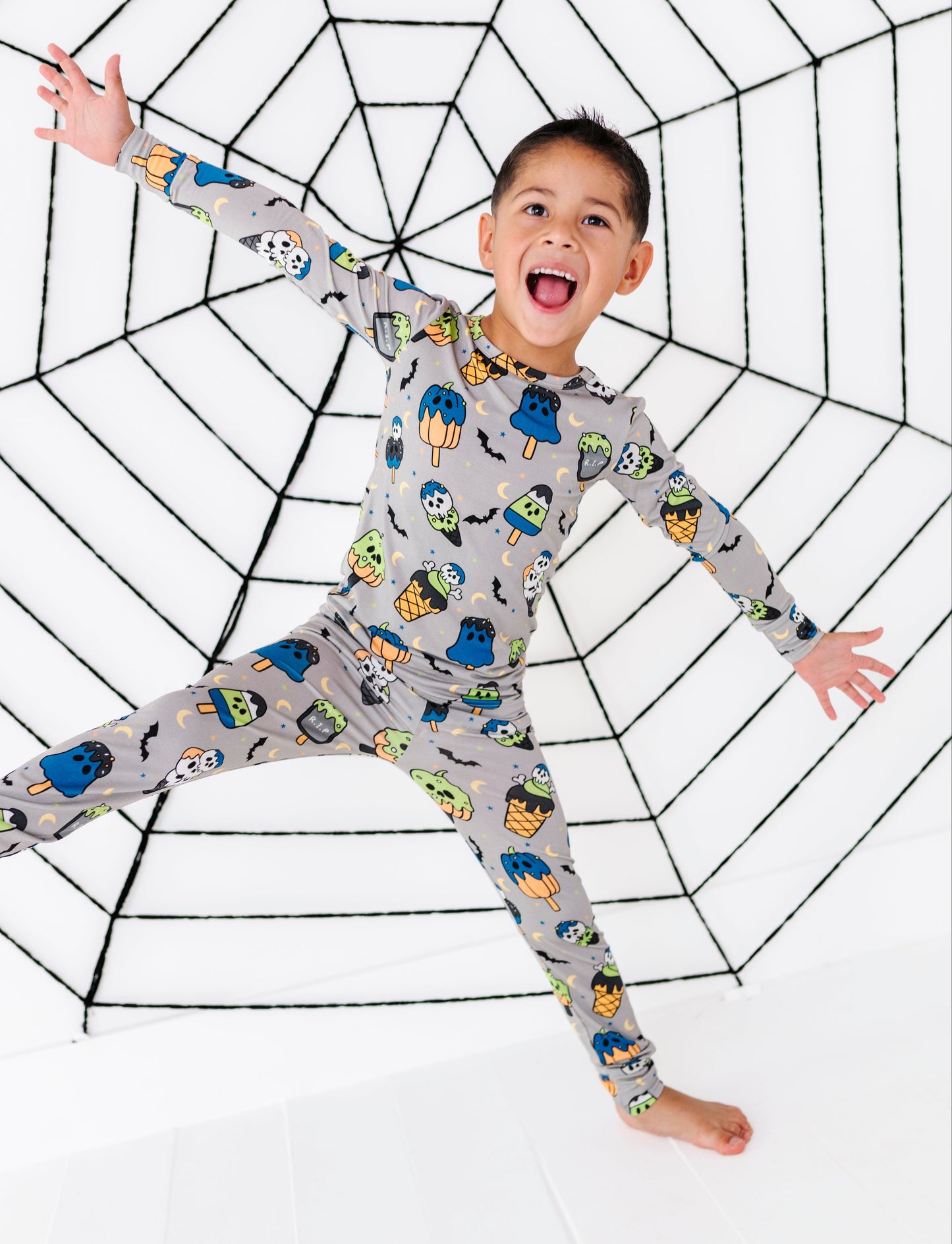 A young boy wearing a gray long-sleeve pajama set with a colorful cartoon monster print, posing with his arms outstretched in front of a large black spiderweb graphic on a white background.