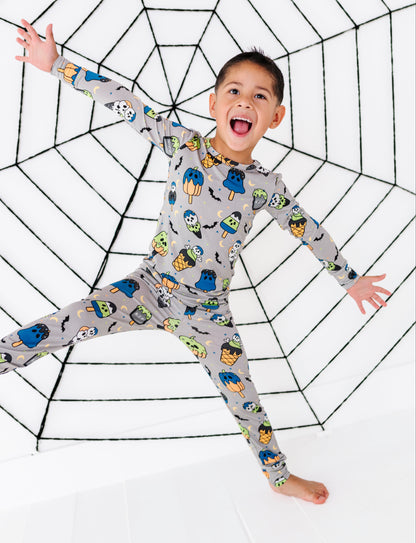 A young boy wearing a gray long-sleeve pajama set with a colorful cartoon monster print, posing with his arms outstretched in front of a large black spiderweb graphic on a white background.