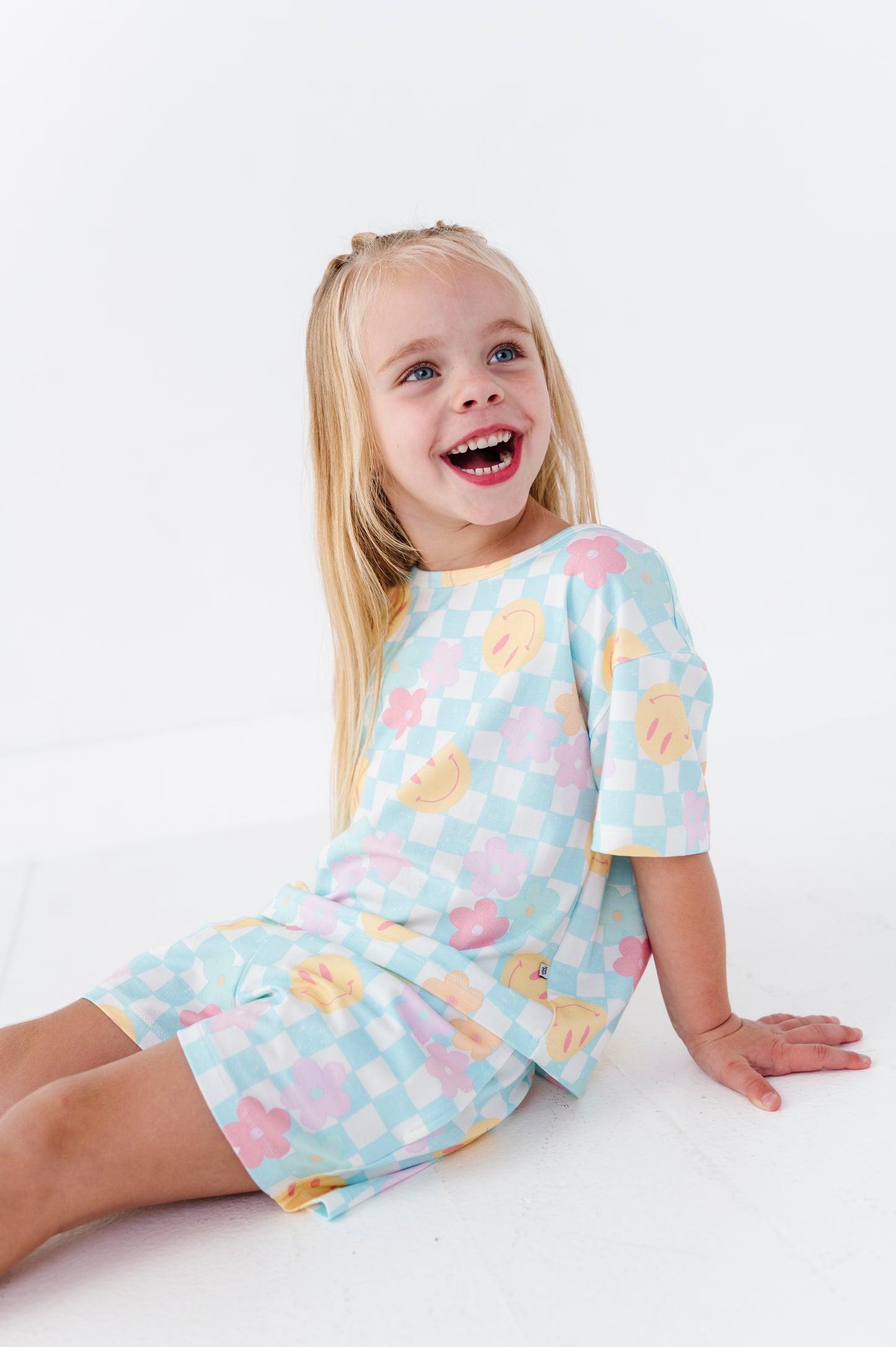 A young blonde girl smiling and laughing while wearing a light blue checkered pajama set with a pastel fruit print, sitting on a white studio floor.