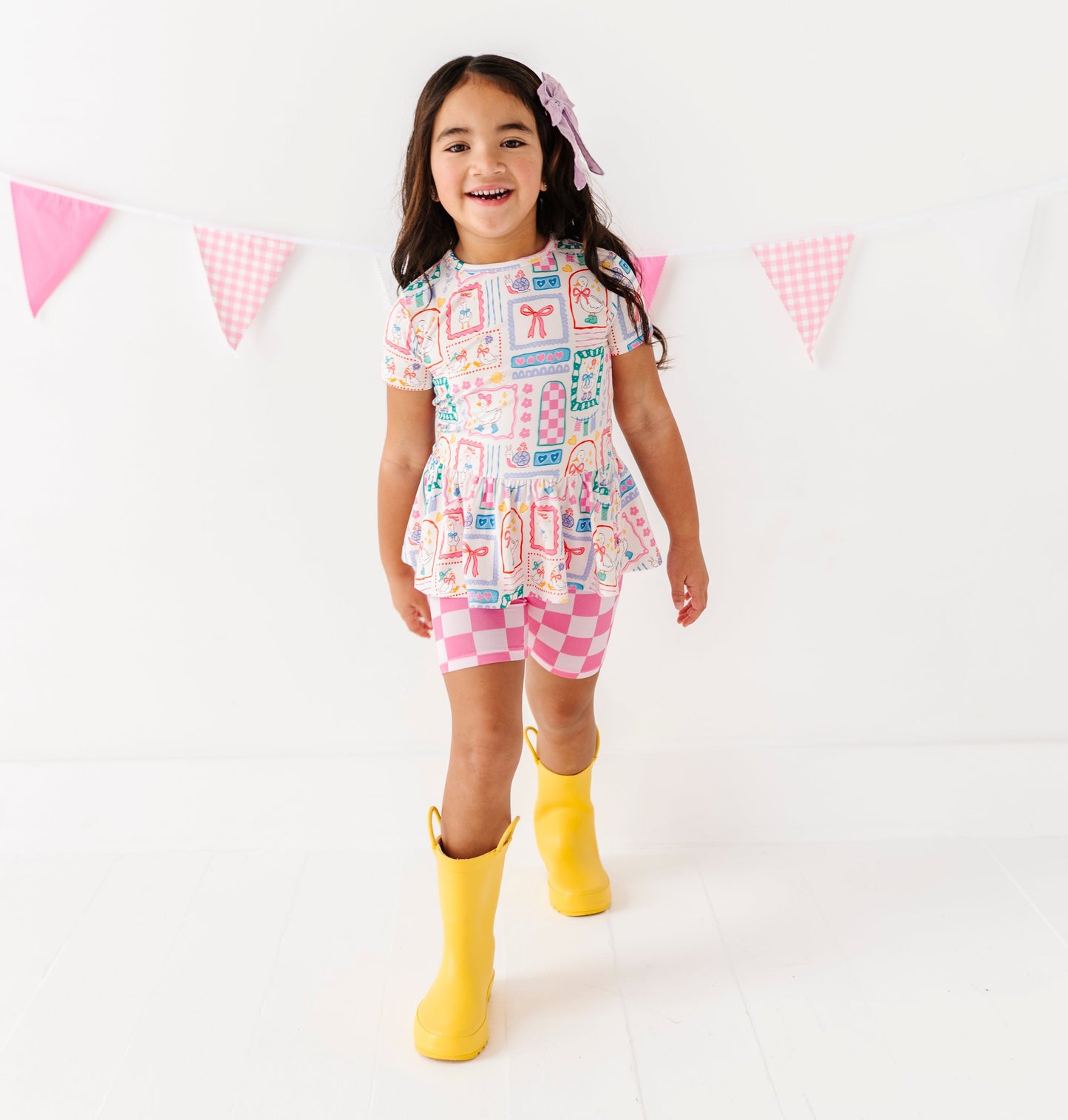 Child wearing Gilly Goose bamboo/cotton peplum top with pink checkered bamboo biker shorts and yellow rain boots against a white background 