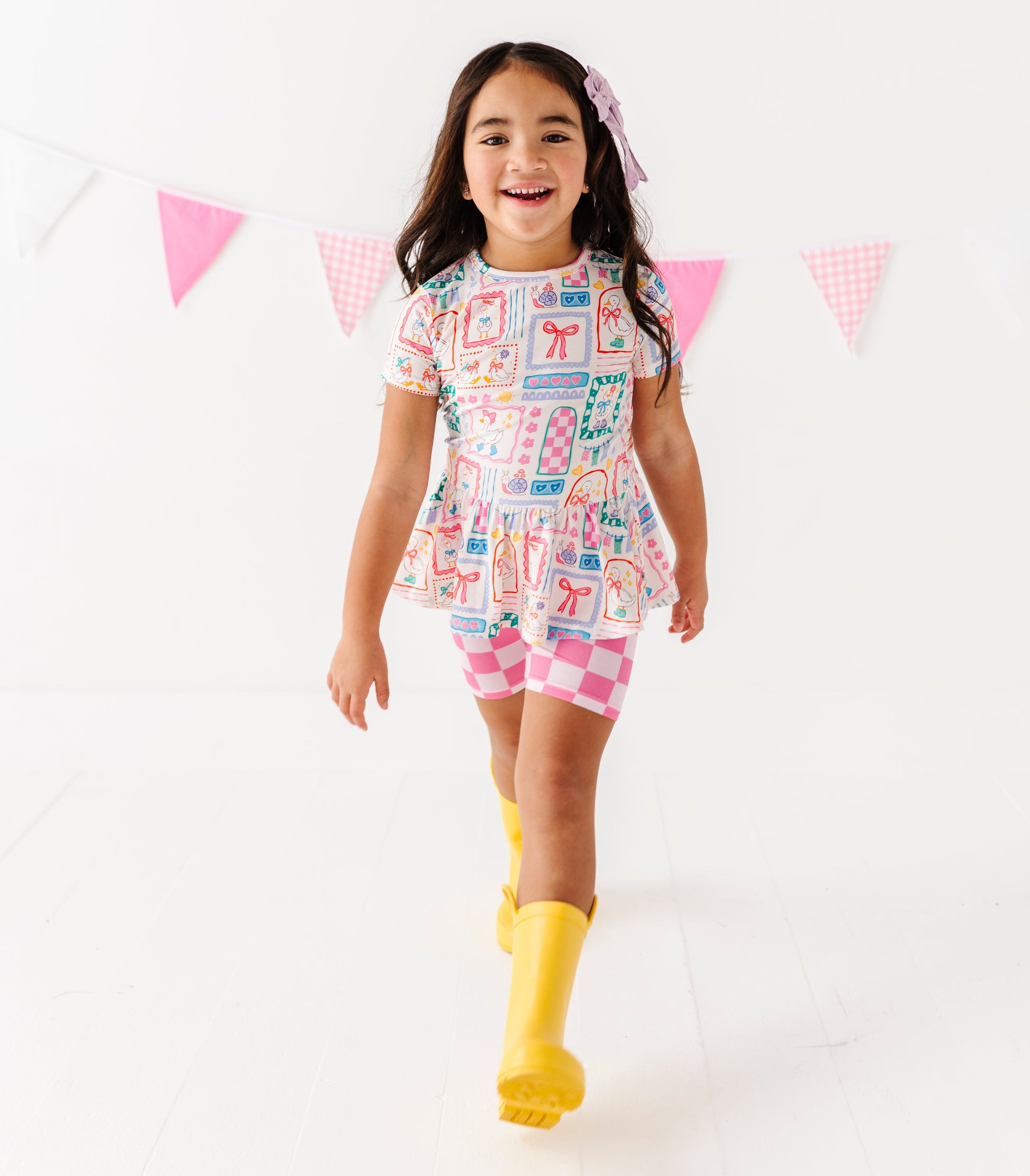 Young girl wearing a Gilly Goose bamboo/cotton blend peplum top with checkered biker shorts and yellow rain boots against a white background 