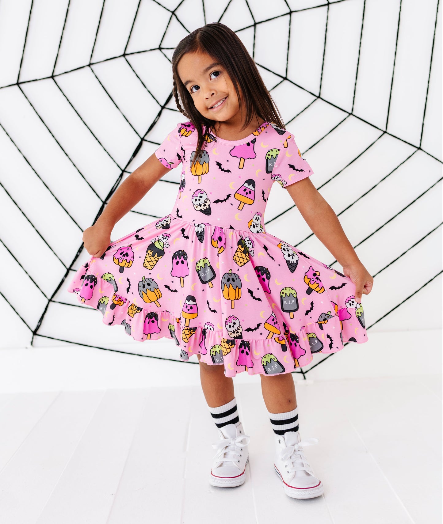 A young girl wearing a pink Halloween-themed dress with a spooky monster print, posing in front of a large black spiderweb prop on a white studio background.