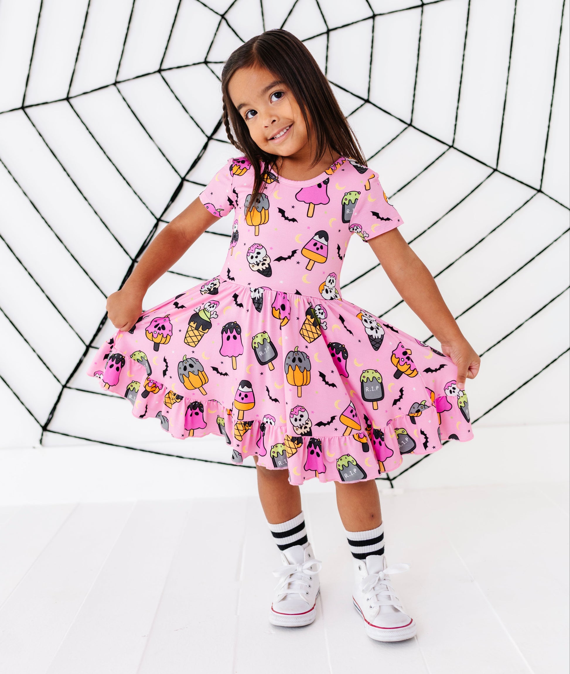 A young girl wearing a pink Halloween-themed dress with a spooky monster print, posing in front of a large black spiderweb prop on a white studio background.