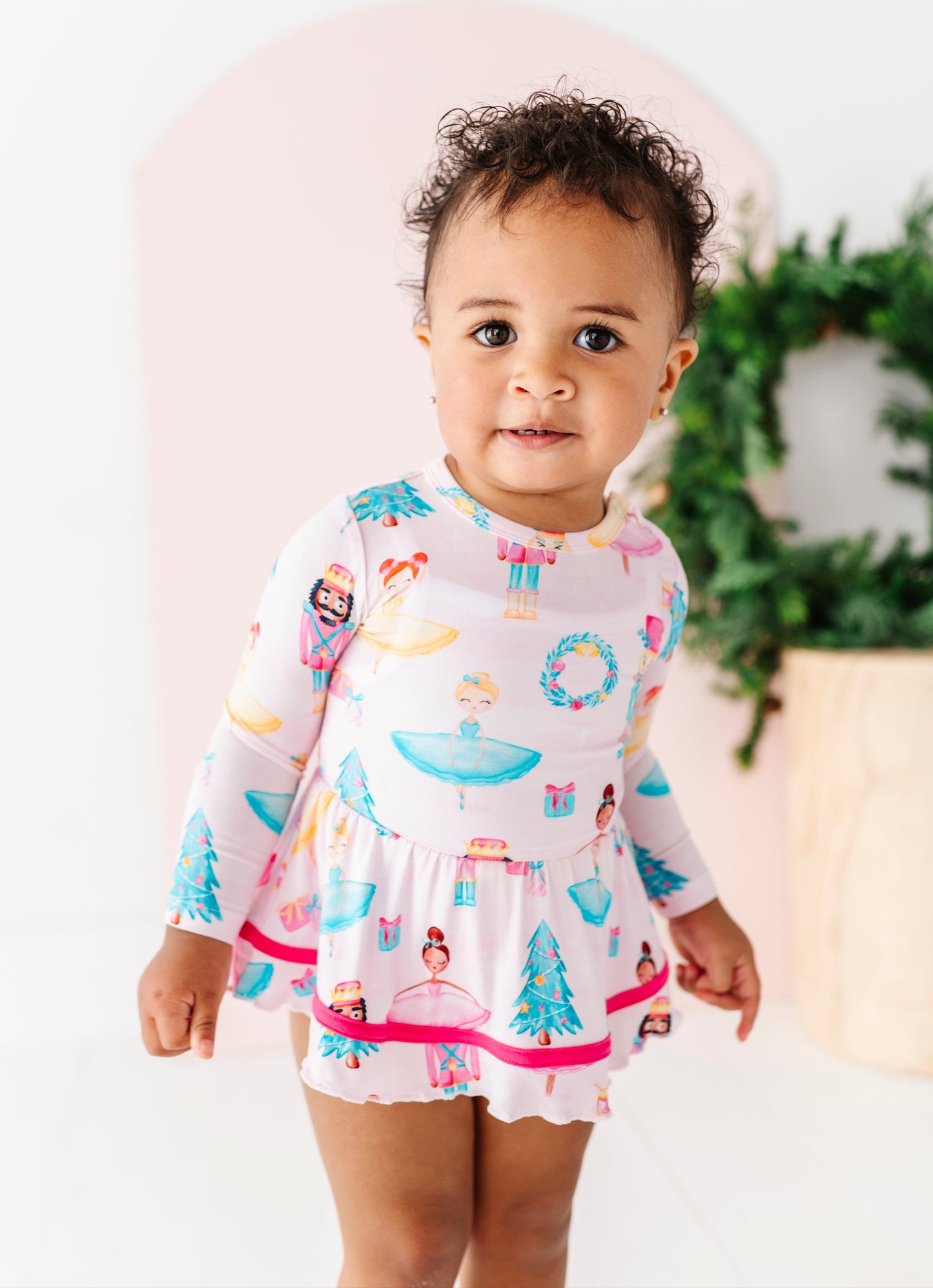 Baby wearing a light pink bamboo long-sleeve twirl dress with a pattern of sugarplum dancers, nutcrackers, presents, Christmas wreaths, and twinkling trees
