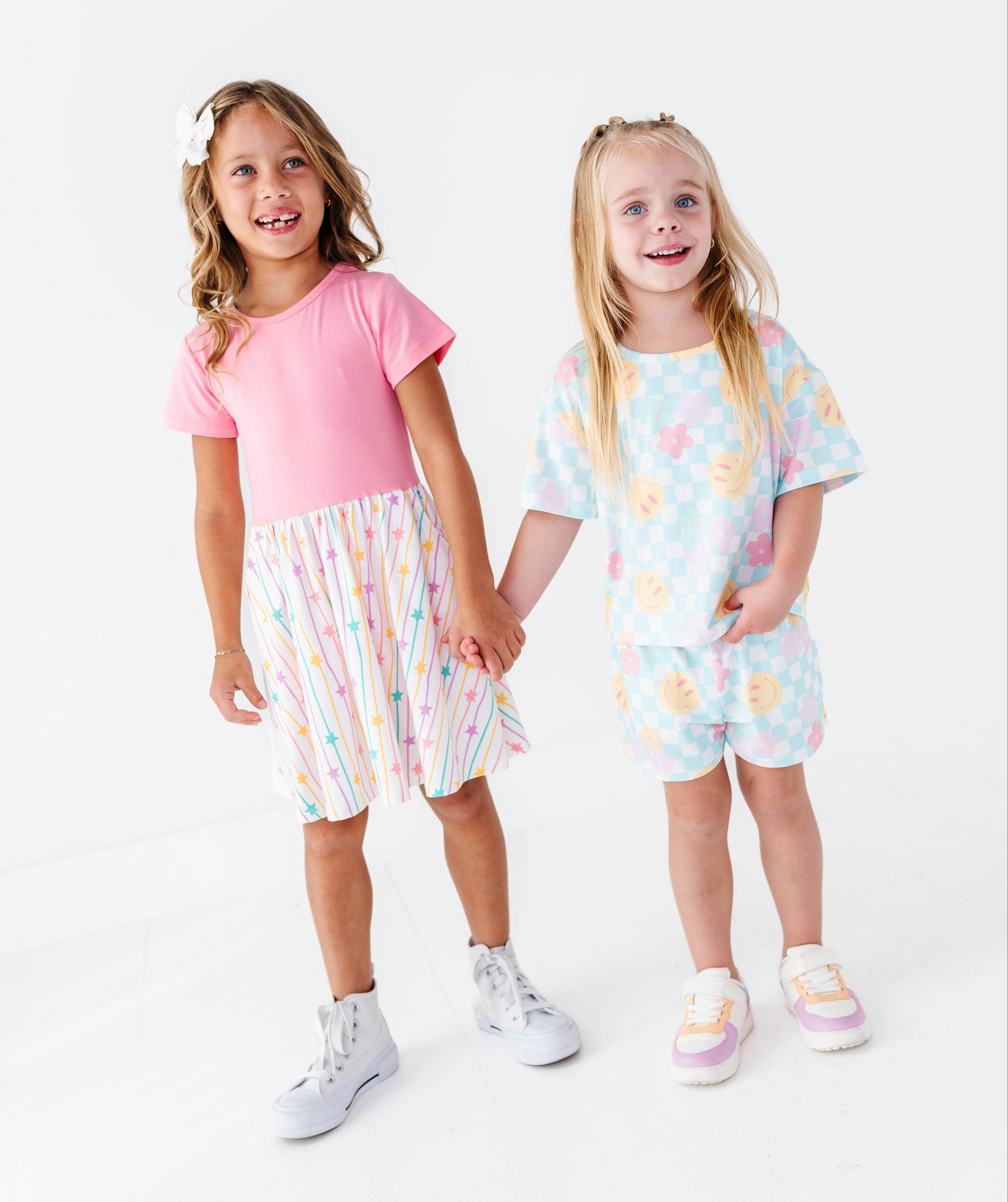 Two young girls standing and holding hands in a studio setting, wearing matching pastel two-piece outfits. The girl on the left wears a pink short-sleeve top with a pastel rainbow-striped skirt, and the girl on the right wears a light blue pastel patterned short-sleeve top and matching shorts.