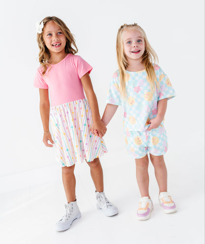 Two young girls standing and holding hands in a studio setting, wearing matching pastel two-piece outfits. The girl on the left wears a pink short-sleeve top with a pastel rainbow-striped skirt, and the girl on the right wears a light blue pastel patterned short-sleeve top and matching shorts.