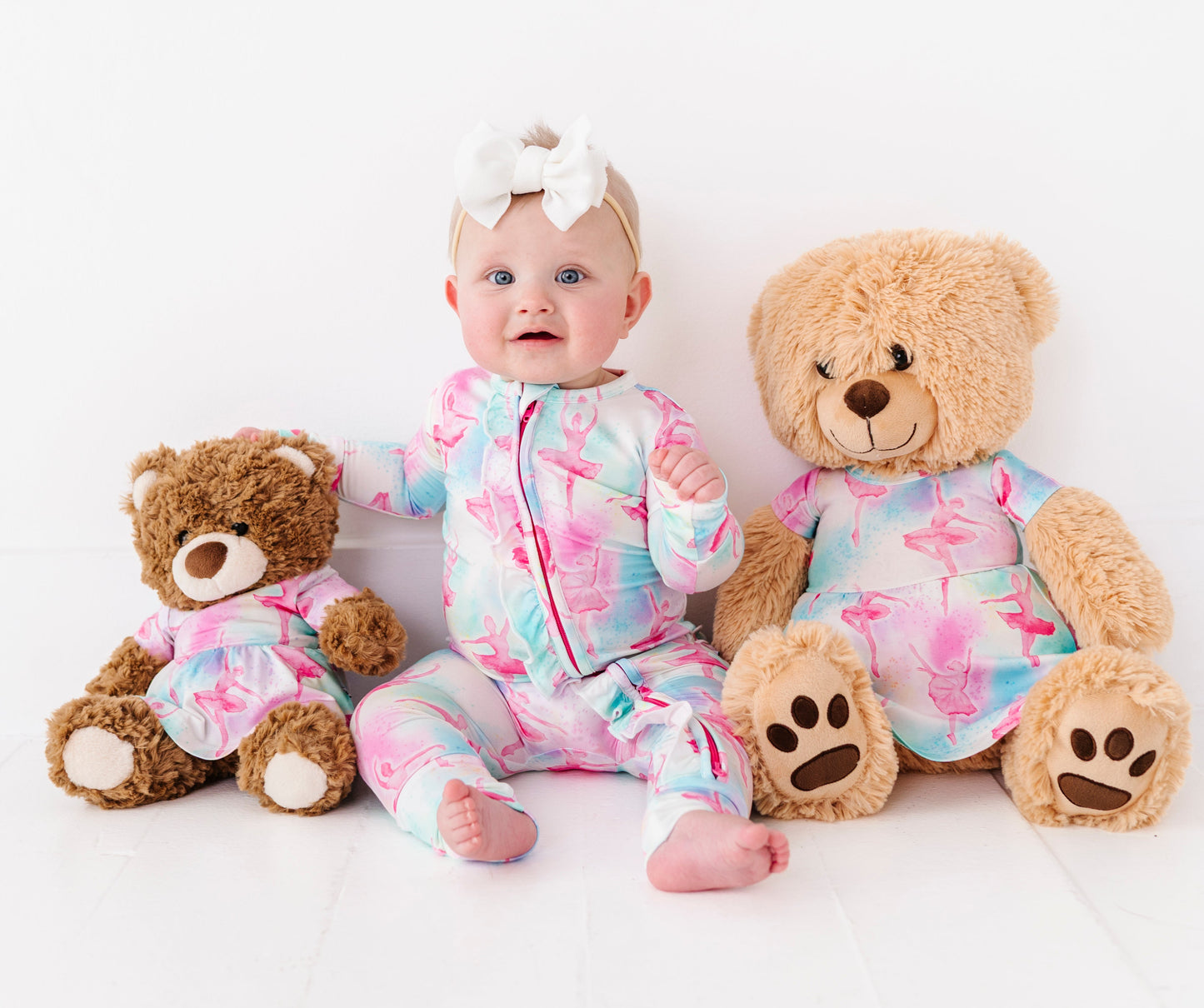 Baby in a bamboo ballet romper sitting between two teddy bears wearing dresses in matching prints on a white background