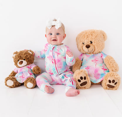 Baby in a ballet themed bamboo zip romper sitting between two teddy bears wearing dresses in a matching ballet pattern on a white background