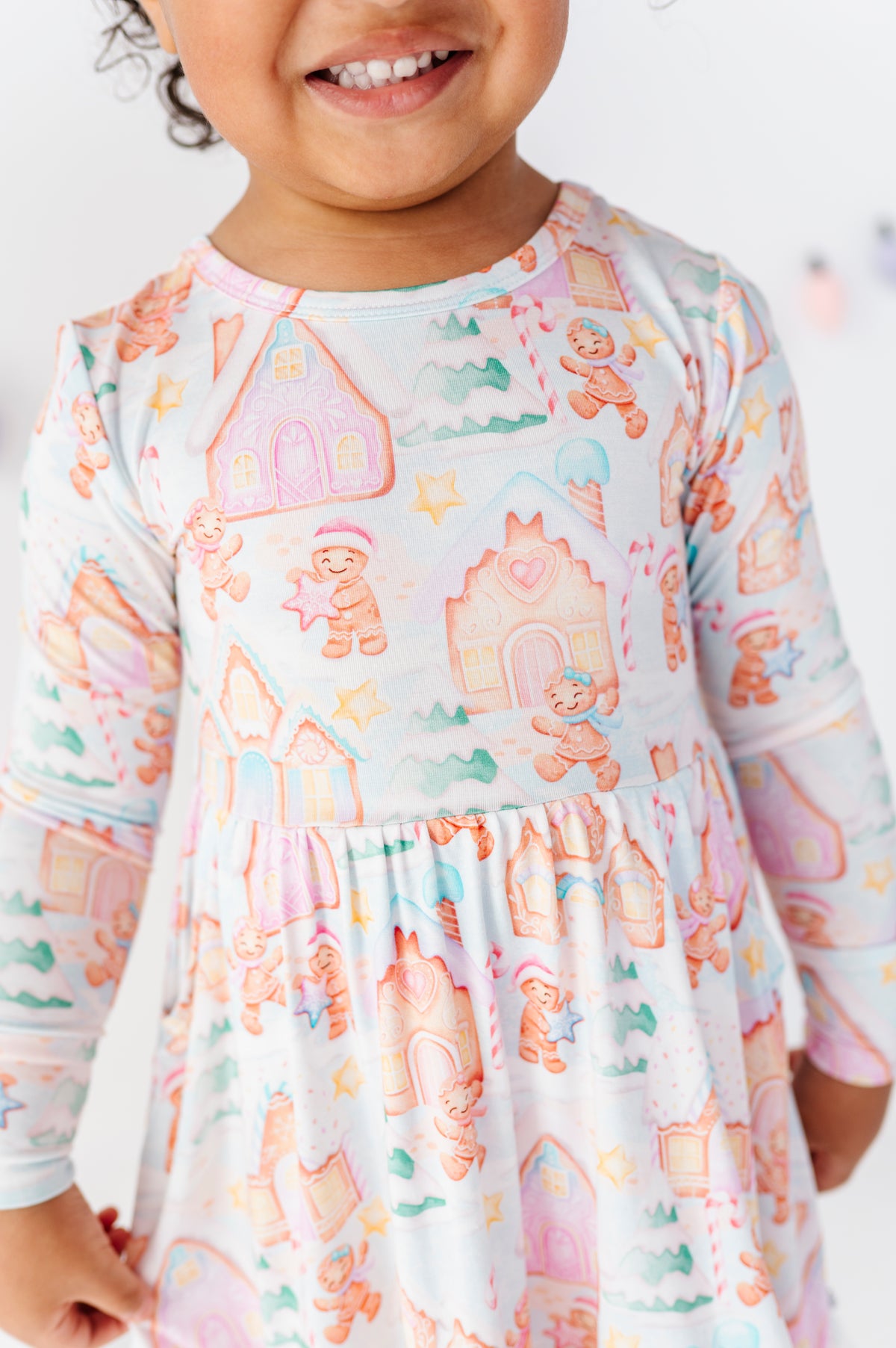 A close-up of a smiling child wearing a long-sleeve dress with a whimsical gingerbread-themed print featuring teddy bears, houses, and stars in pastel pink, orange, and blue on a white background.