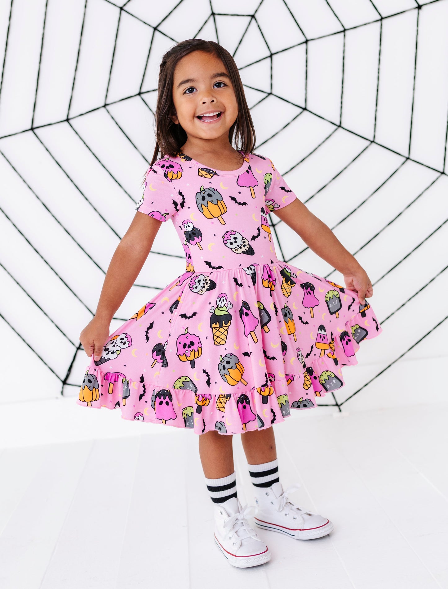 A young girl stands in a studio wearing a pink short-sleeve dress covered in a colorful Halloween print of pumpkins, ghosts, bats, and mummies. She is holding out the sides of the skirt to show its volume, with white socks and white sneakers, against a white background with a black spiderweb graphic.