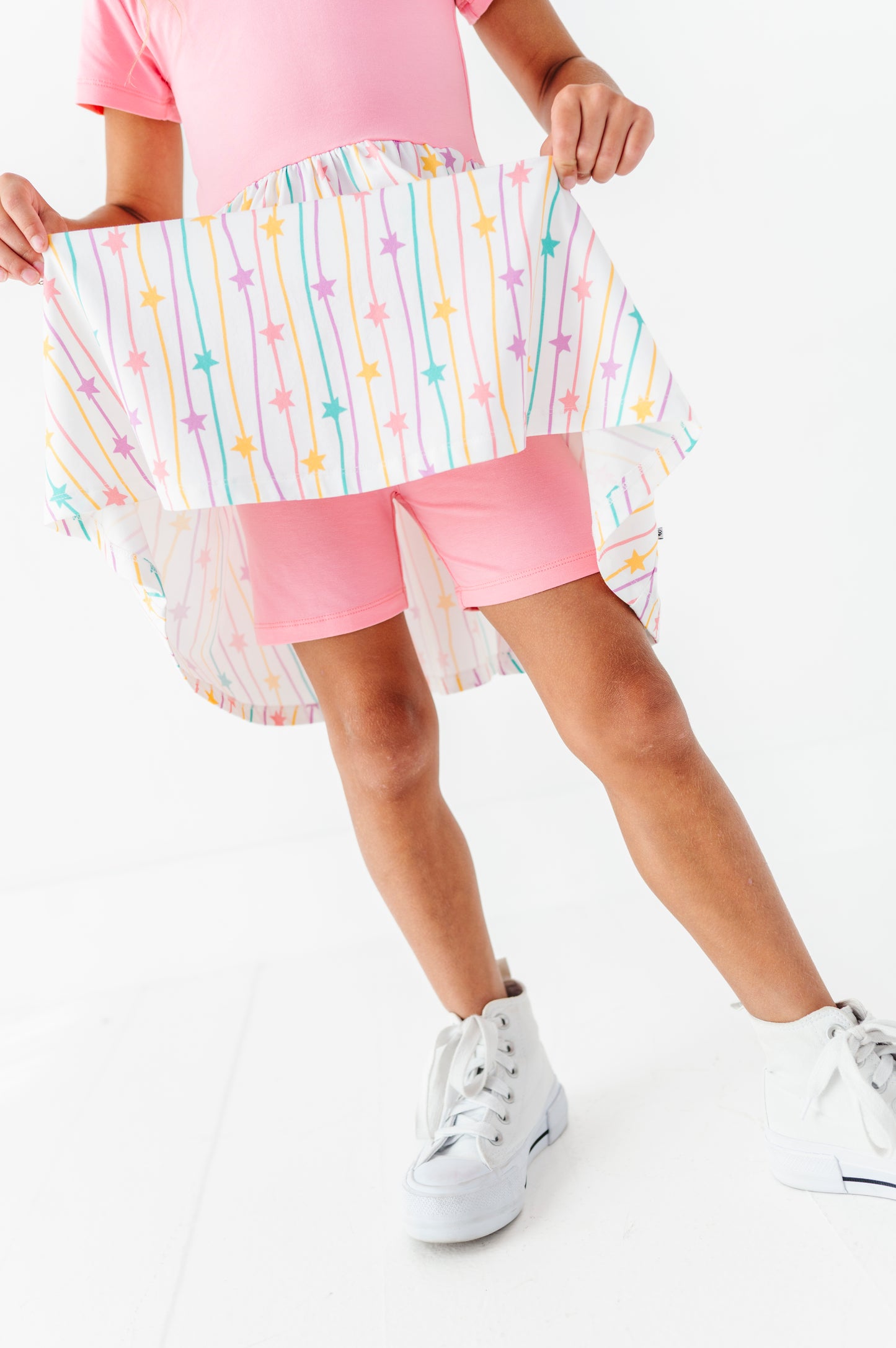 A child wearing a pink outfit and white sneakers holds up a white high-low skirt with pastel rainbow star and stripe patterns against a white studio background.