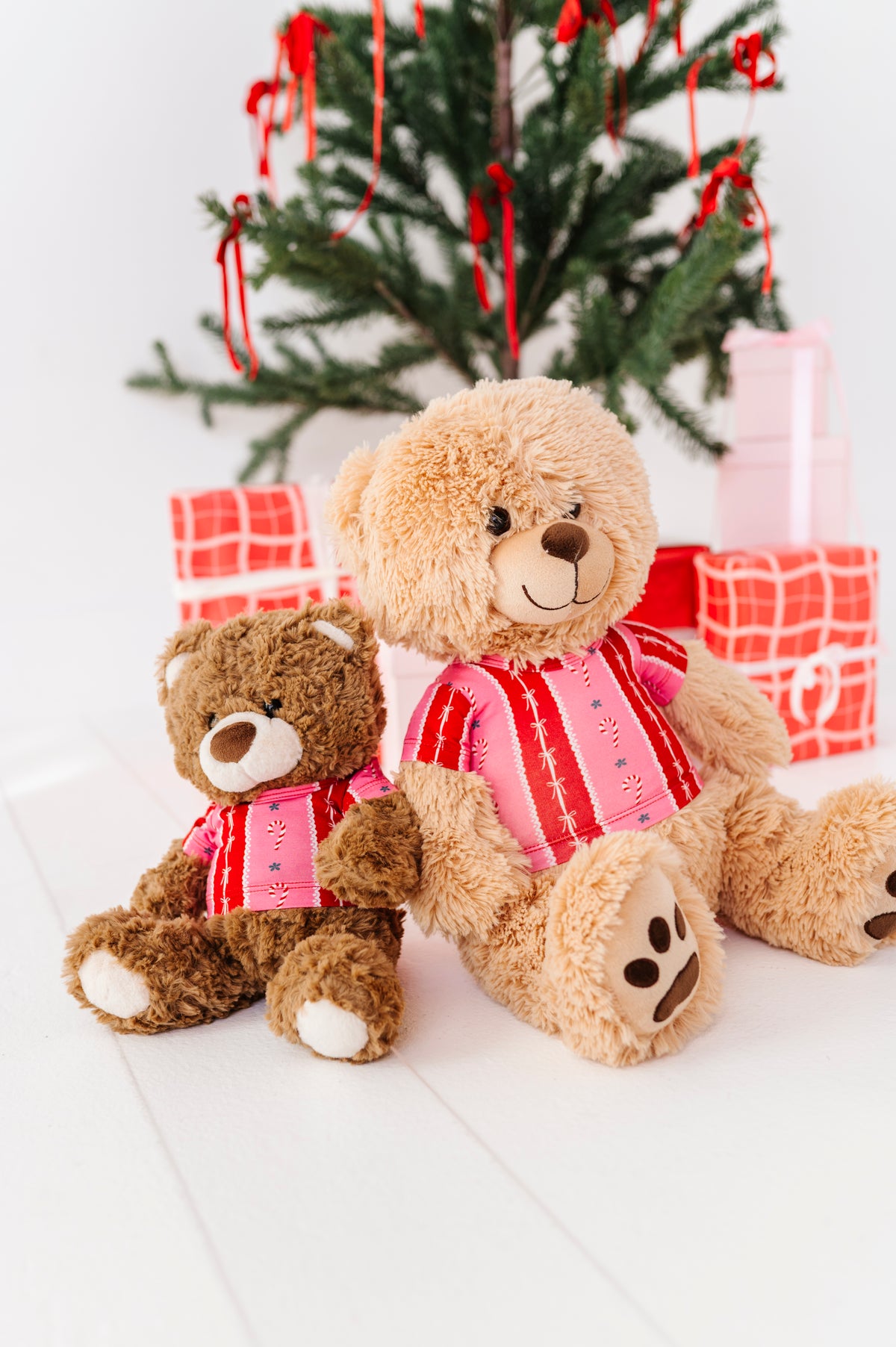 Two plush teddy bears sitting together on a white surface, with a decorated Christmas tree and wrapped gifts in the background. The larger bear is tan and wears a pink striped shirt, while the smaller bear is brown and wears a matching pink striped shirt.