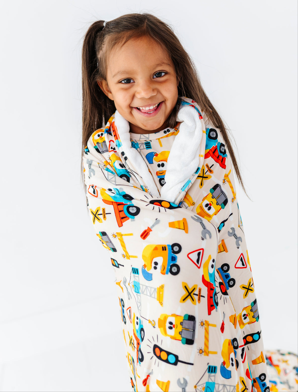 A young girl with dark hair in a ponytail smiling while wrapped in a cozy fleece blanket featuring a colorful construction worker and truck pattern on a white background.
