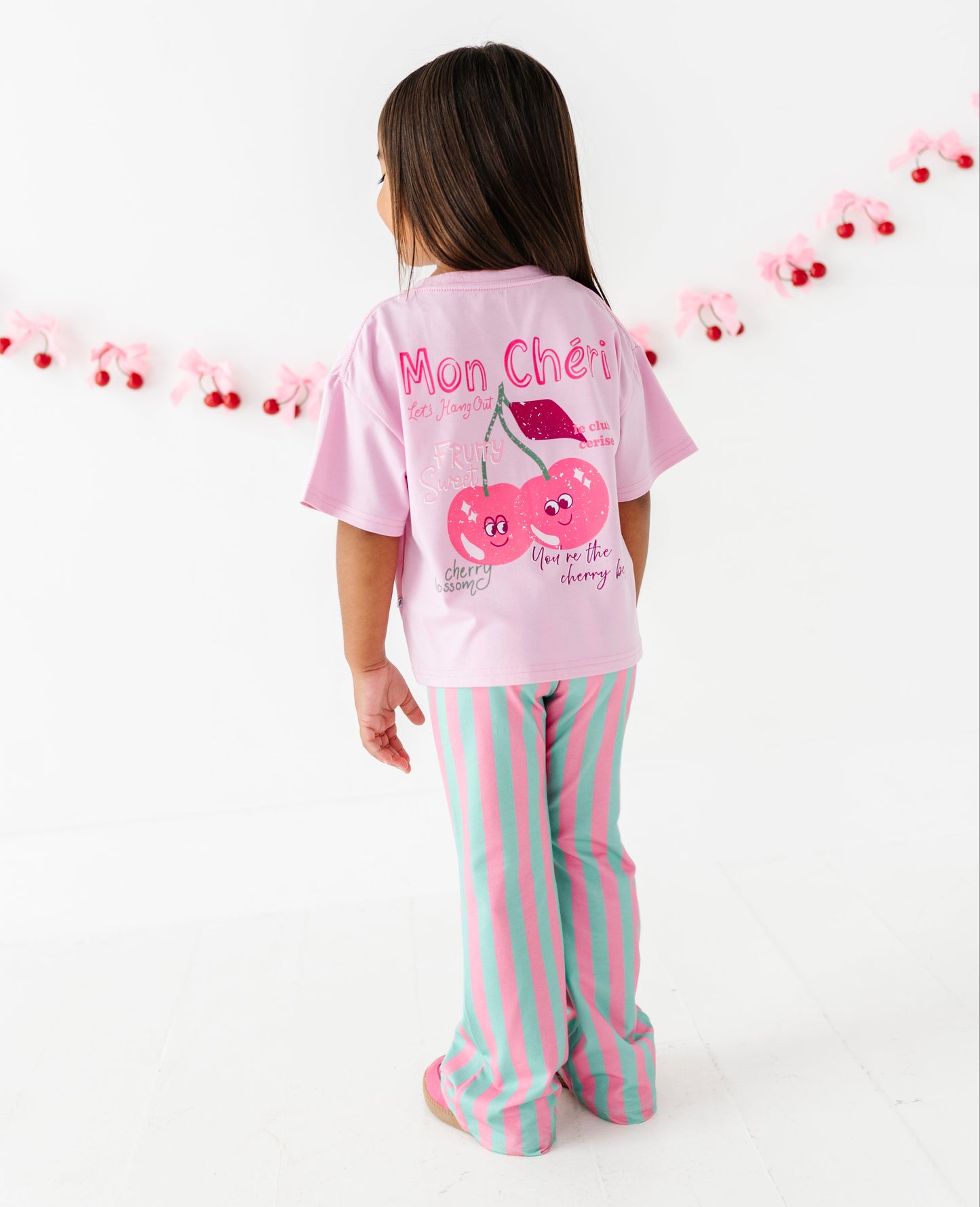 A young girl viewed from behind wearing a pink t-shirt with a cherry graphic and the text 'Mon Cheri' and pink and mint green striped pants, standing against a white studio background with a cherry garland.