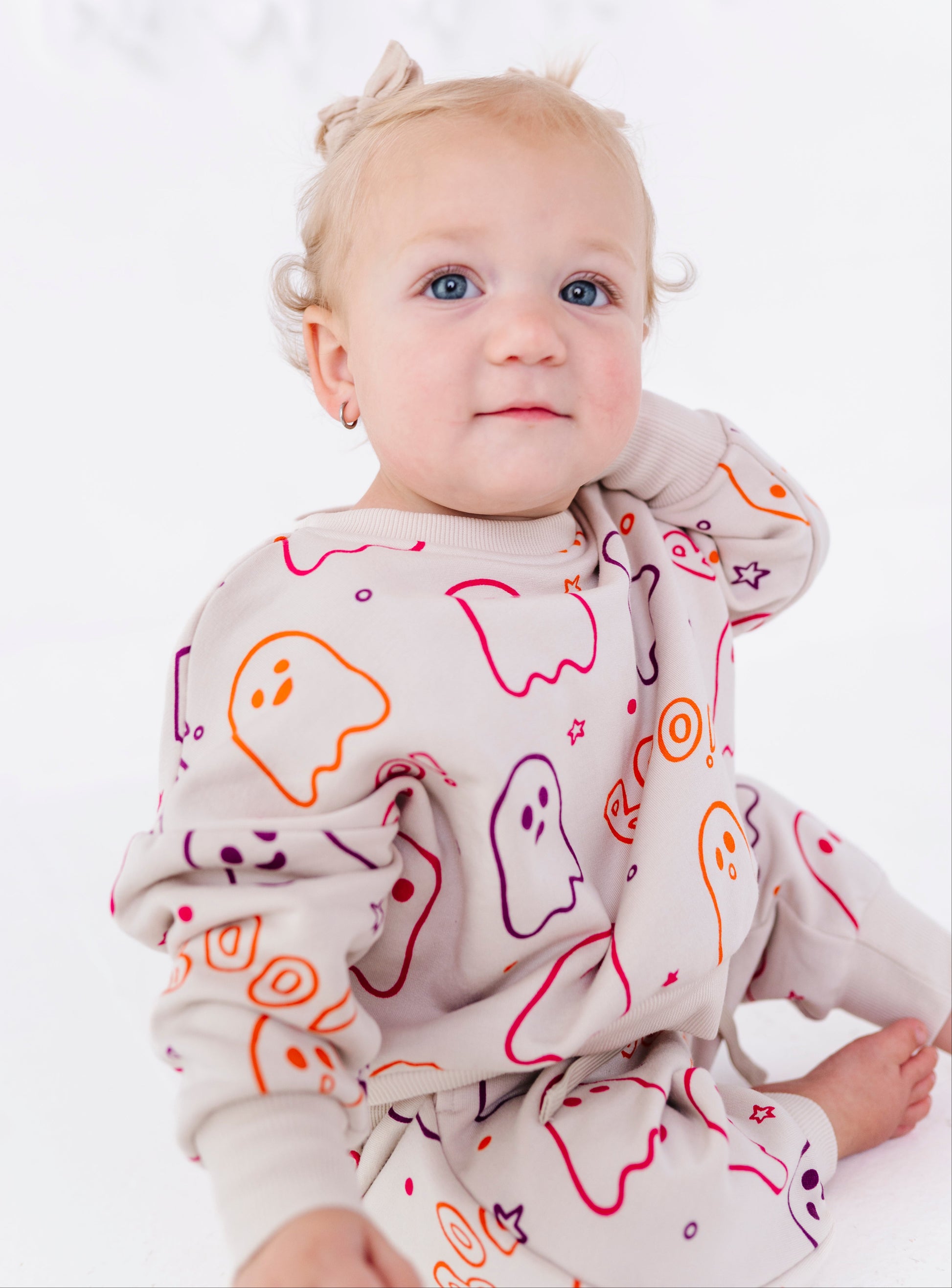 A baby wearing a light beige long-sleeve pajama set with a colorful ghost pattern, sitting against a white background.