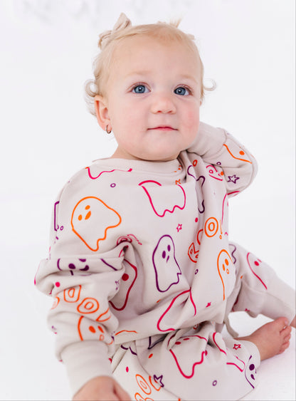 A baby wearing a light beige long-sleeve pajama set with a colorful ghost pattern, sitting against a white background.