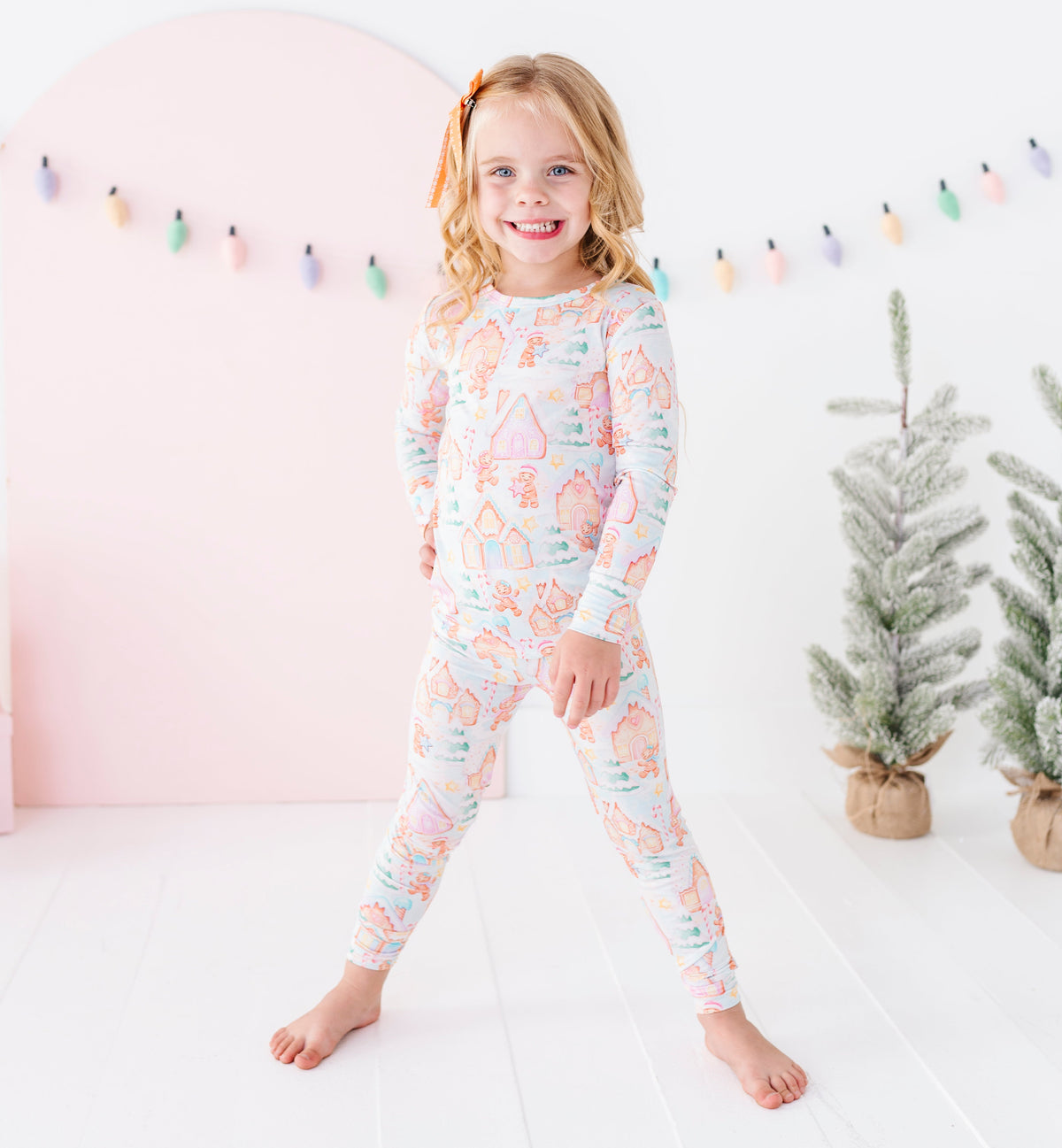 A young girl with blonde hair stands barefoot in a bright white room wearing a long-sleeve two-piece bamboo pajama set with a pastel gingerbread village print. The background features a soft pink arch, colorful hanging string lights, and two small potted artificial trees.