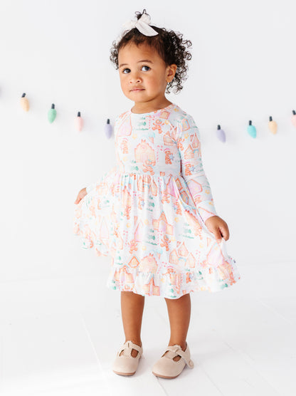 A toddler girl wearing a long-sleeve ruffled dress with a pastel multicolor fairy print, standing against a white studio background with a colorful string of fairy lights.