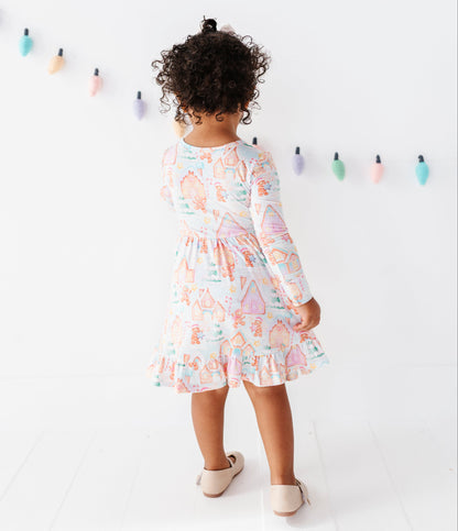 A toddler girl with curly dark hair is seen from behind wearing a long-sleeve ruffled dress with a pastel multicolor print. She is standing in a bright white room with a string of colorful hanging light bulbs on the wall.