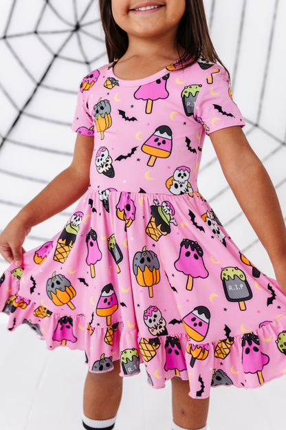 A young girl wearing a pink short-sleeve skater dress with a Halloween-themed print of popsicles, bats, and tombstones, shown against a white background with a black spiderweb graphic.