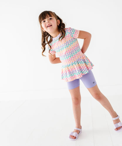 Young girl wearing a colorful bamboo/cotton top and light purple bamboo biker shorts on a white background