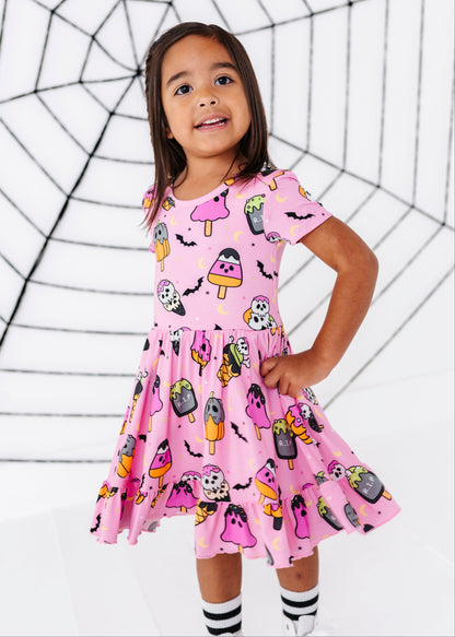 A young girl wearing a pink short-sleeve dress with a Halloween-themed print of ghosts, bats, and popsicles, standing in front of a white background with a black spiderweb graphic.