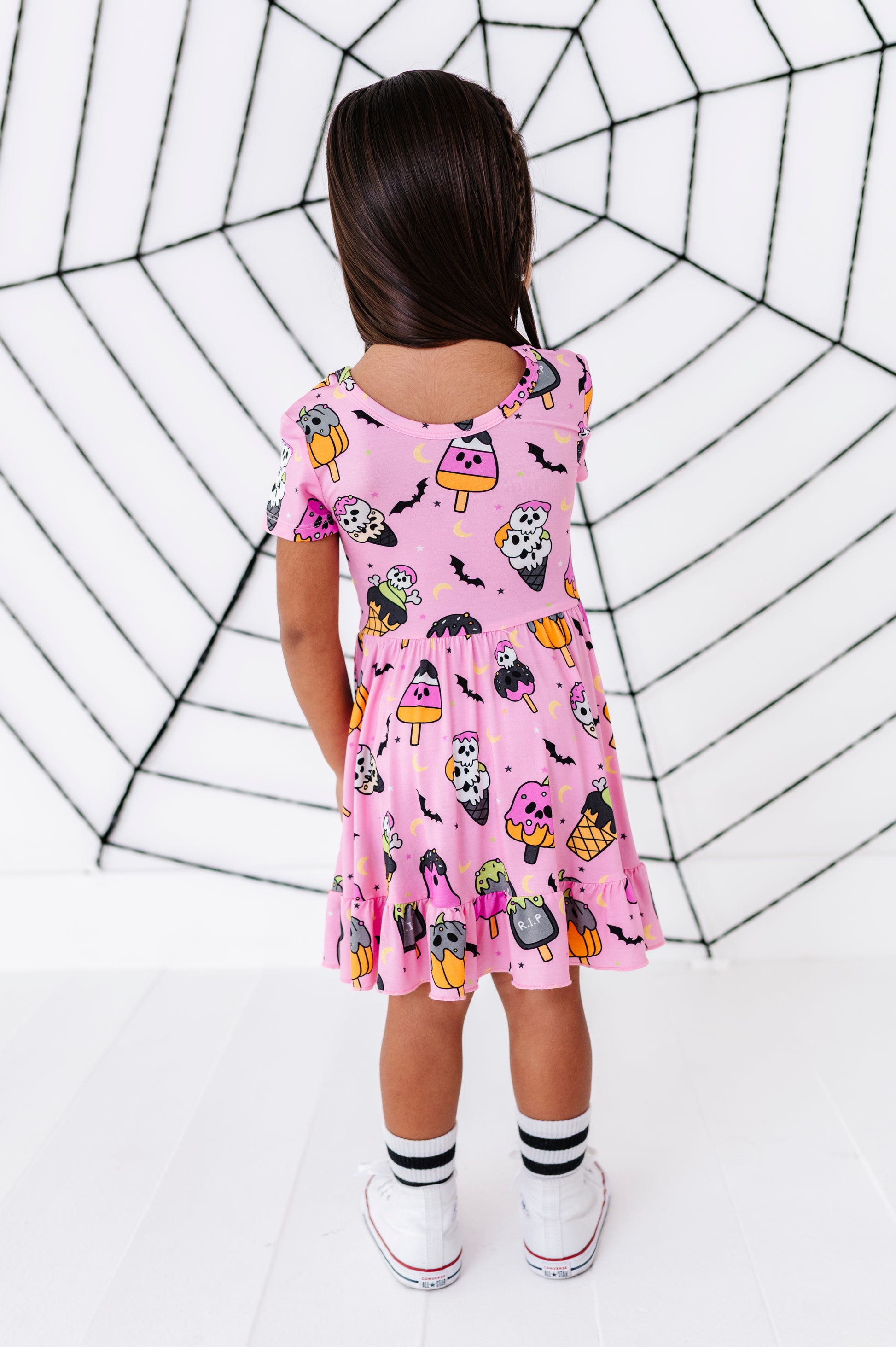 A young girl viewed from behind wearing a pink short-sleeve dress with a Halloween print of ghosts, bats, pumpkins, and cauldrons, standing in front of a white wall with a black spiderweb pattern.