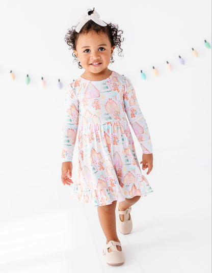 A toddler girl wearing a long-sleeve pastel dress with a whimsical cartoon print, walking forward in a bright white studio setting with a bow headband and beige shoes.