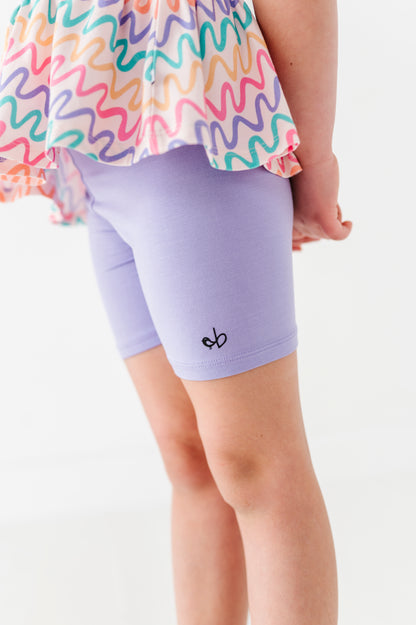 Little girl wearing a colorful bamboo/cotton top and purple bamboo biker shorts on a white background