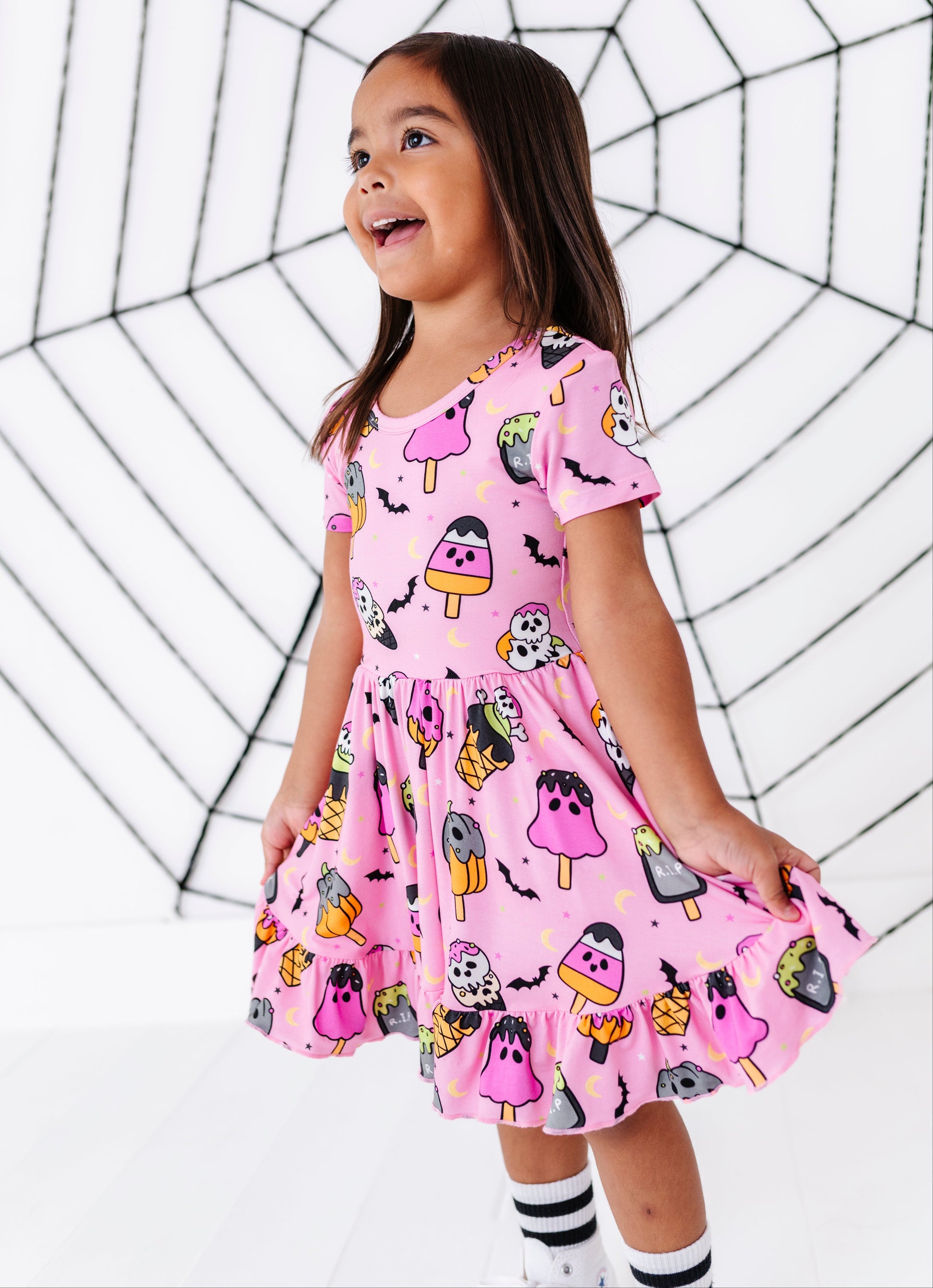 A young girl with long brown hair stands in a white studio wearing a pink short-sleeve dress with a Halloween-themed print of ghosts, pumpkins, bats, and popsicles. She is holding out the skirt of the dress, and a black spiderweb graphic is printed on the background.