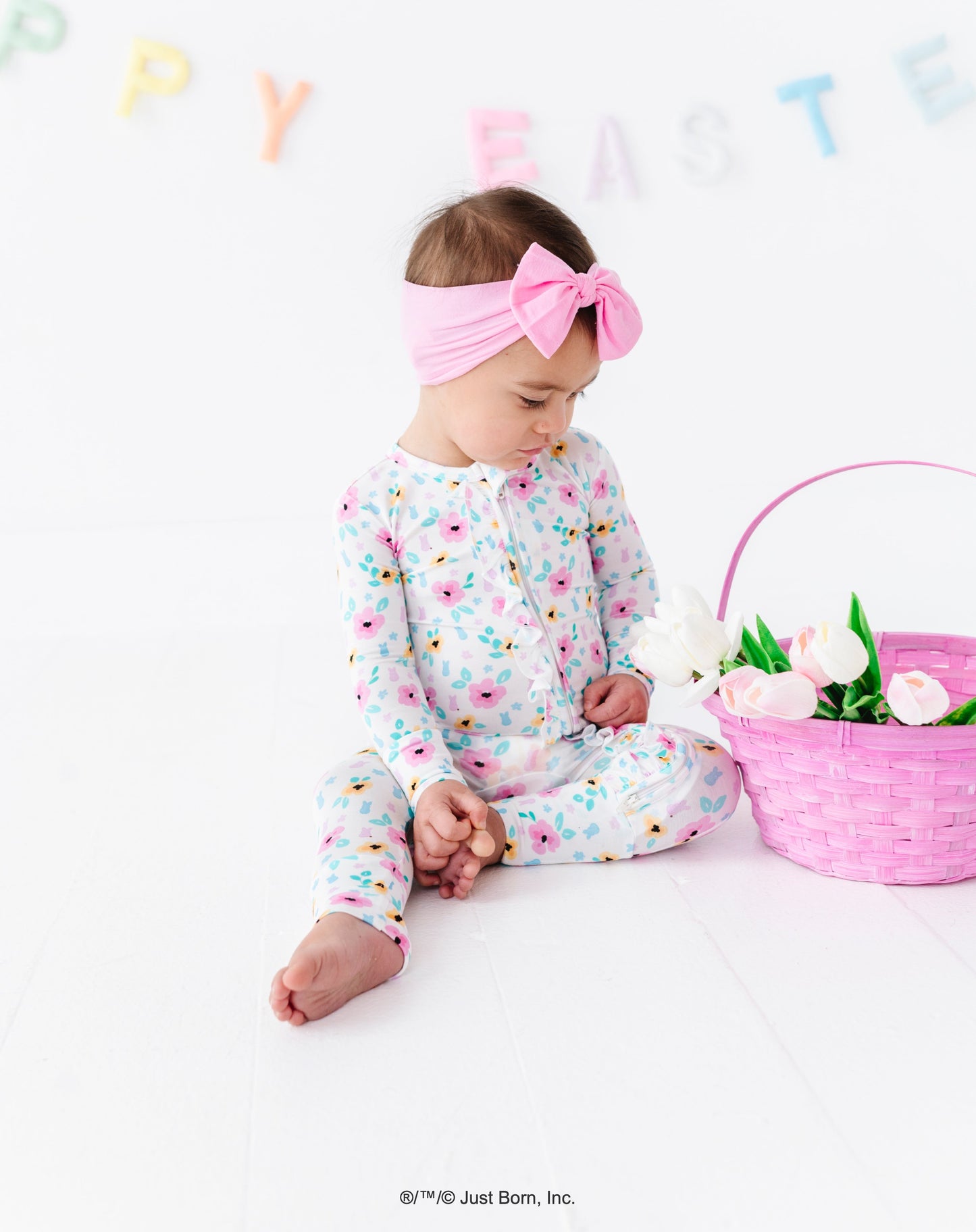 Baby in floral PEEPS® romper with pink Easter basket and tulips, sitting on a white floor with 'Happy Easter' banner.
