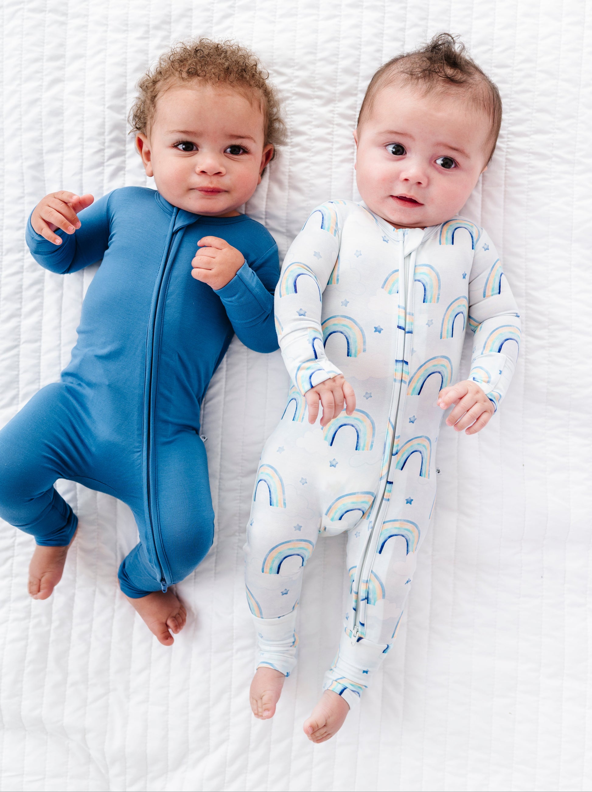 Two babies wearing blue and rainbow themed bamboo zip rompers on a white textured surface