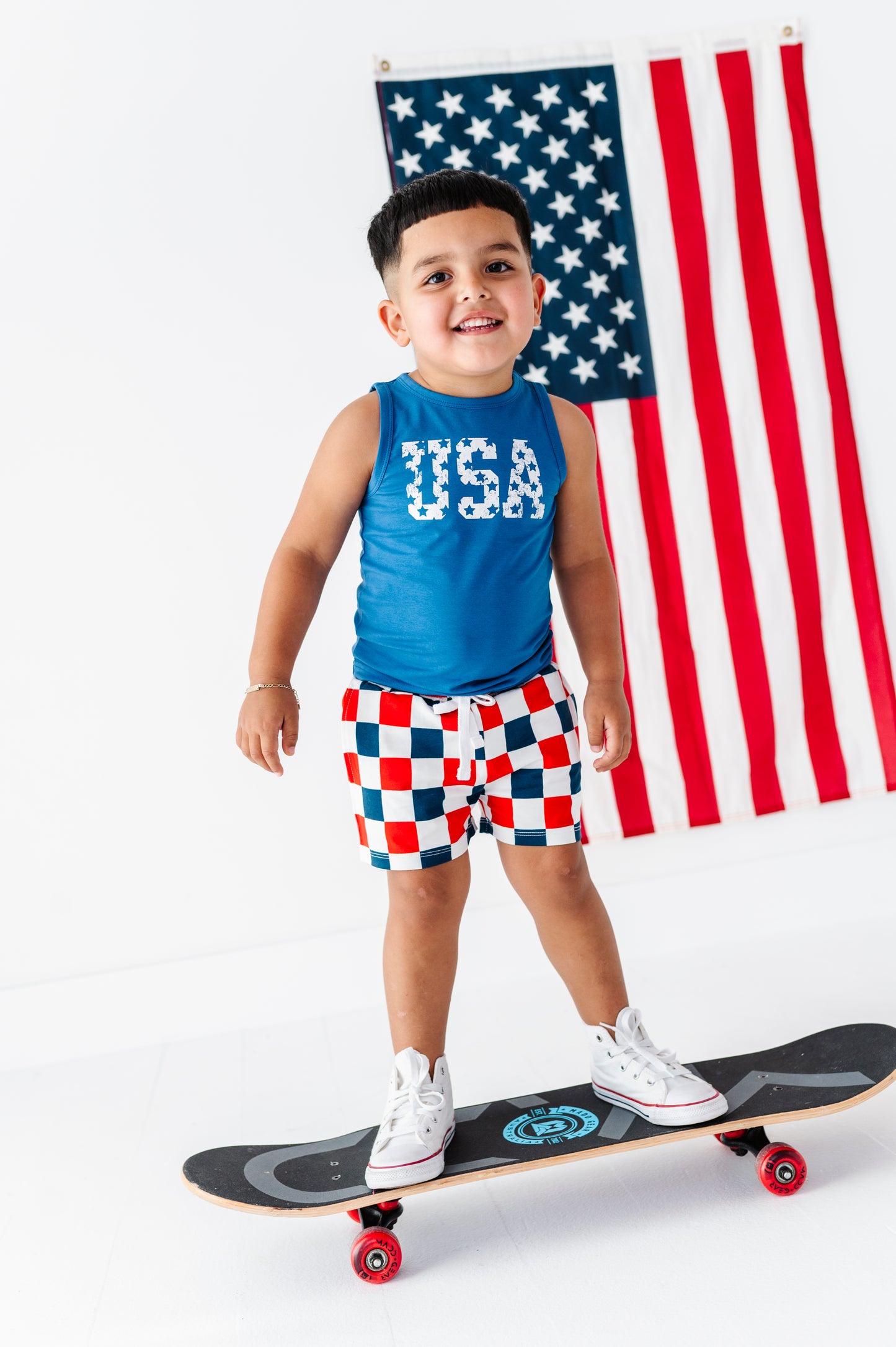 Child wearing a patriotic tank top and standing on a skateboard with an American flag in the background