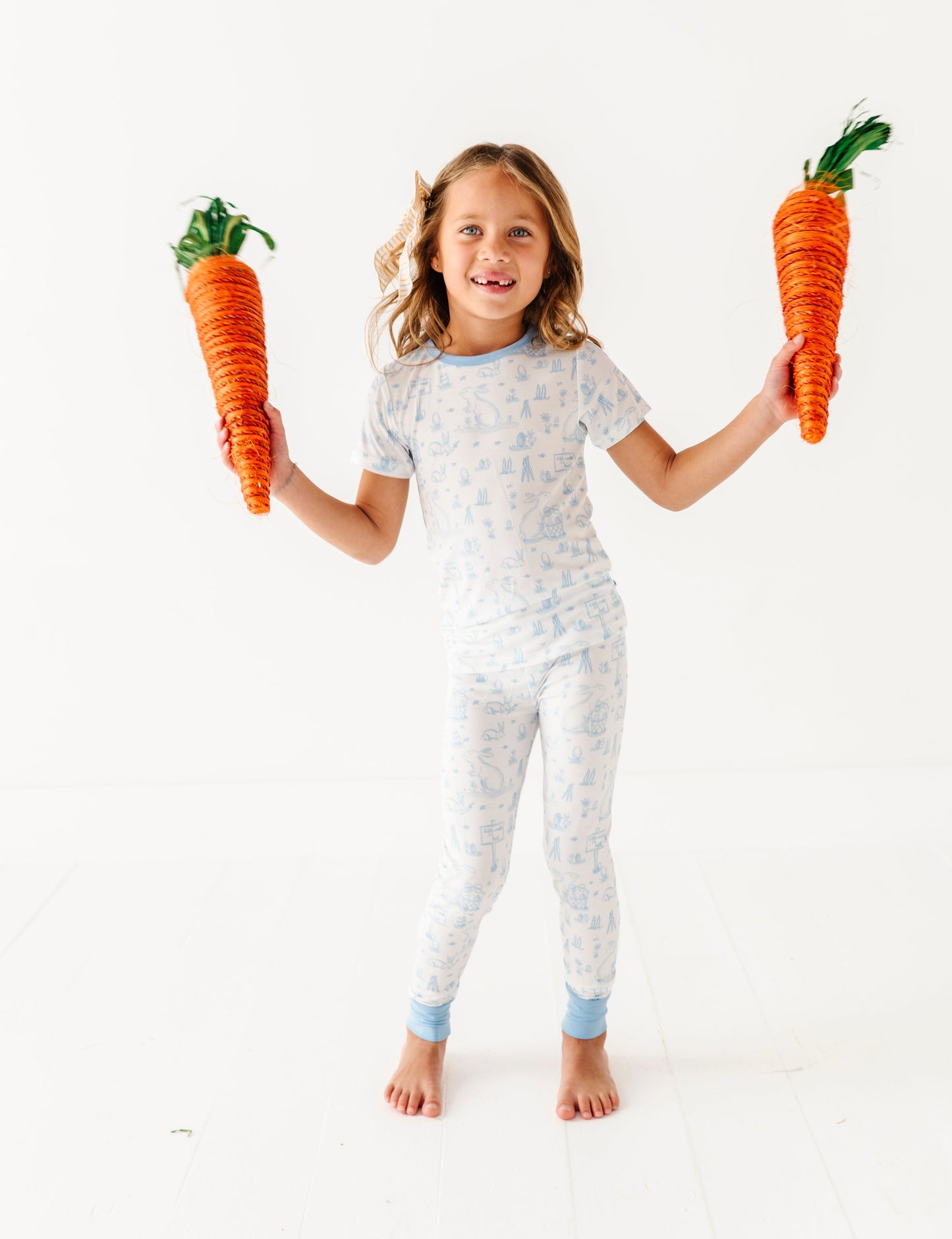 Child wearing a blue and white patterned Easter pajamas holding two large toy carrots.