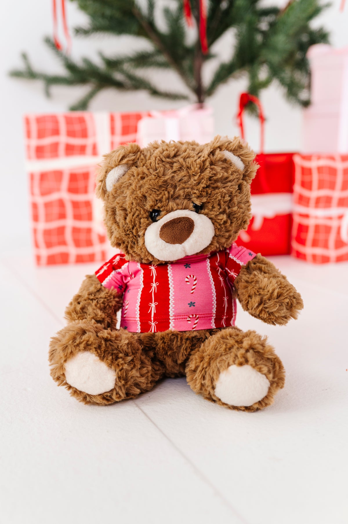 A brown plush teddy bear wearing a pink and red striped bamboo shirt with candy cane details, sitting in front of wrapped Christmas presents and a blurred Christmas tree.