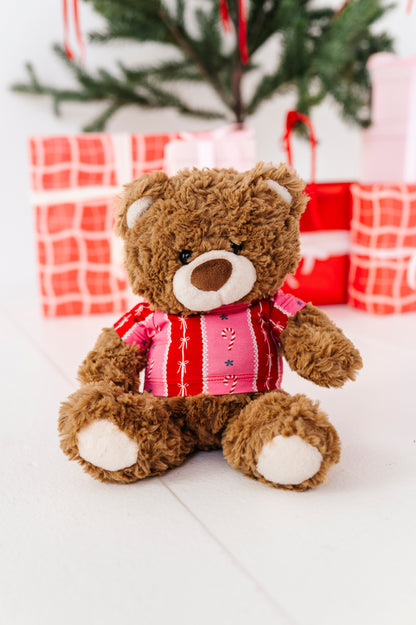 A brown plush teddy bear wearing a pink and red striped bamboo shirt with candy cane details, sitting in front of wrapped Christmas presents and a blurred Christmas tree.