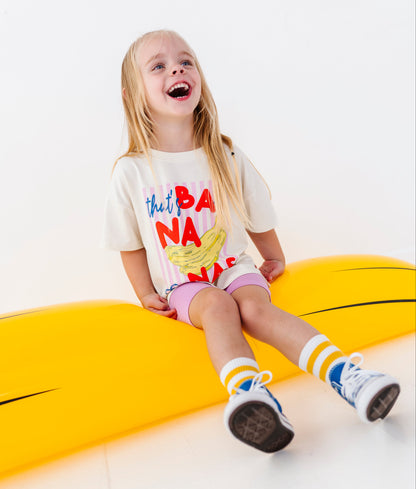 A young girl with blonde hair sits smiling on a large yellow banana-shaped slide against a clean white studio background. She is wearing a white graphic T-shirt with colorful text and a banana illustration, pink shorts, striped socks, and blue sneakers.